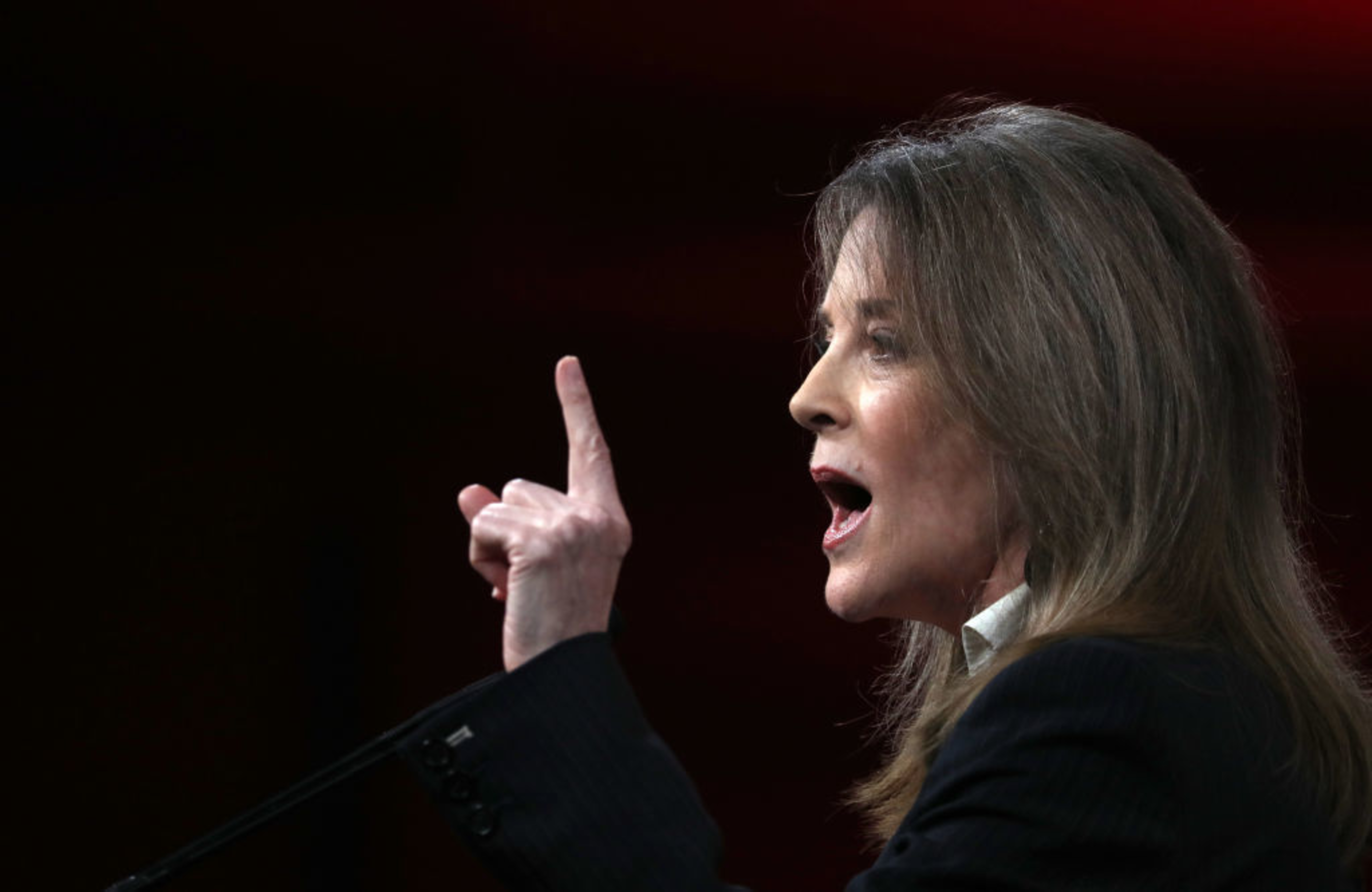 A woman passionately speaking at a podium, raising her index finger, with a blurred red background.