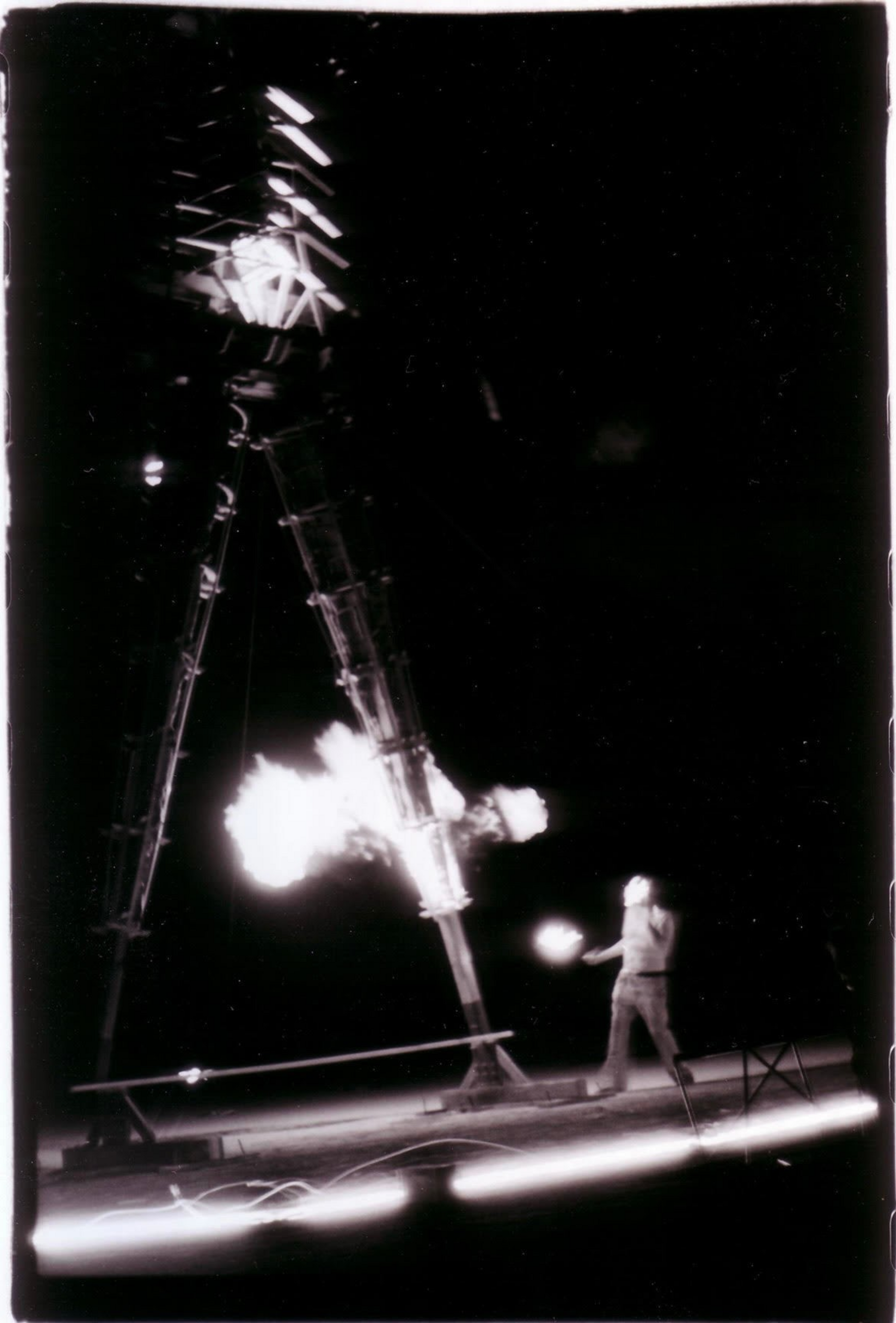 A large mechanical figure made of metal is walking on stilts, while a person nearby appears to be holding a flame in a dark setting.
