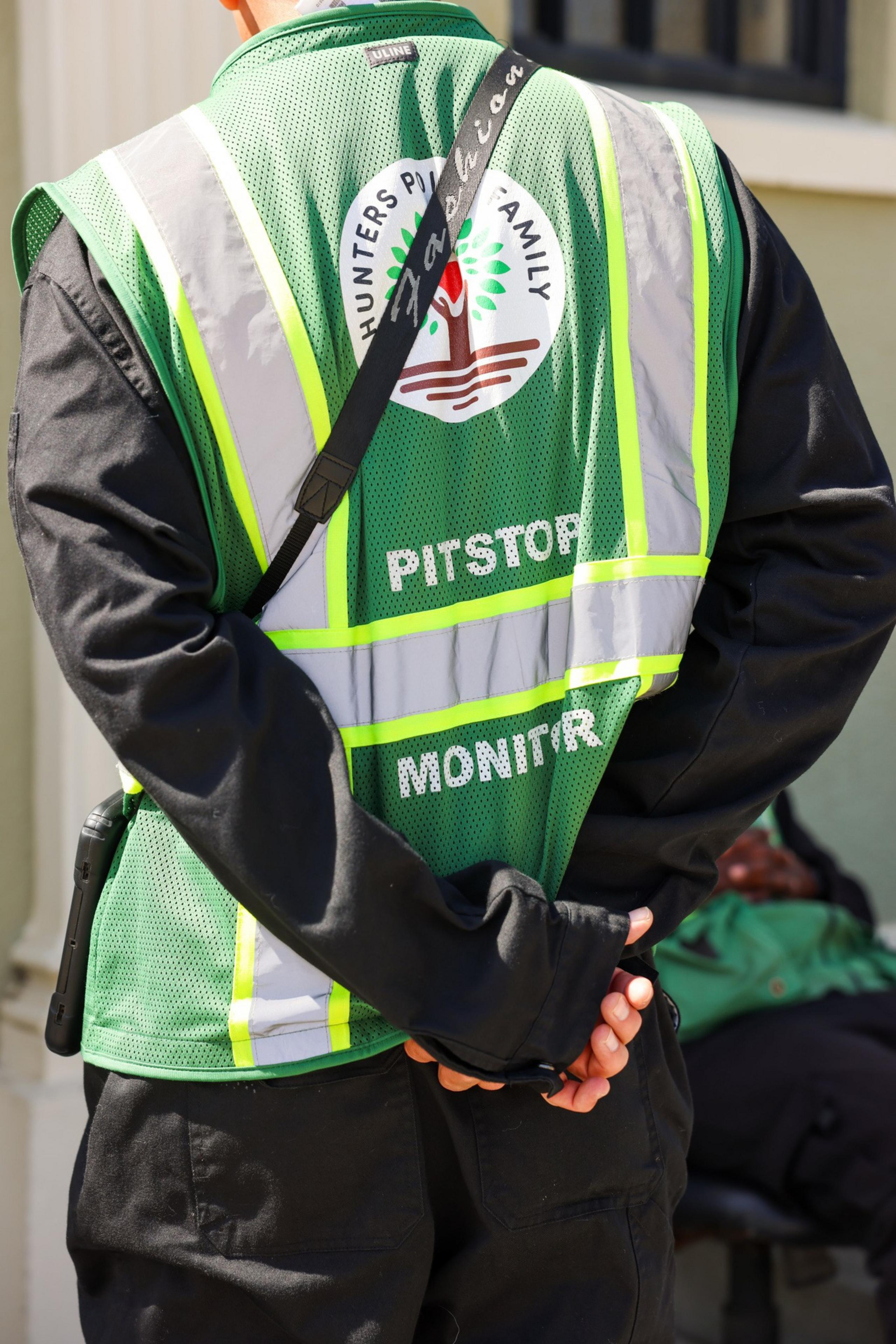 A person wearing a green safety vest with reflective strips stands with their hands clasped behind their back. The vest reads &quot;Hunters Point Family&quot; and &quot;Pitstop Monitor.&quot;
