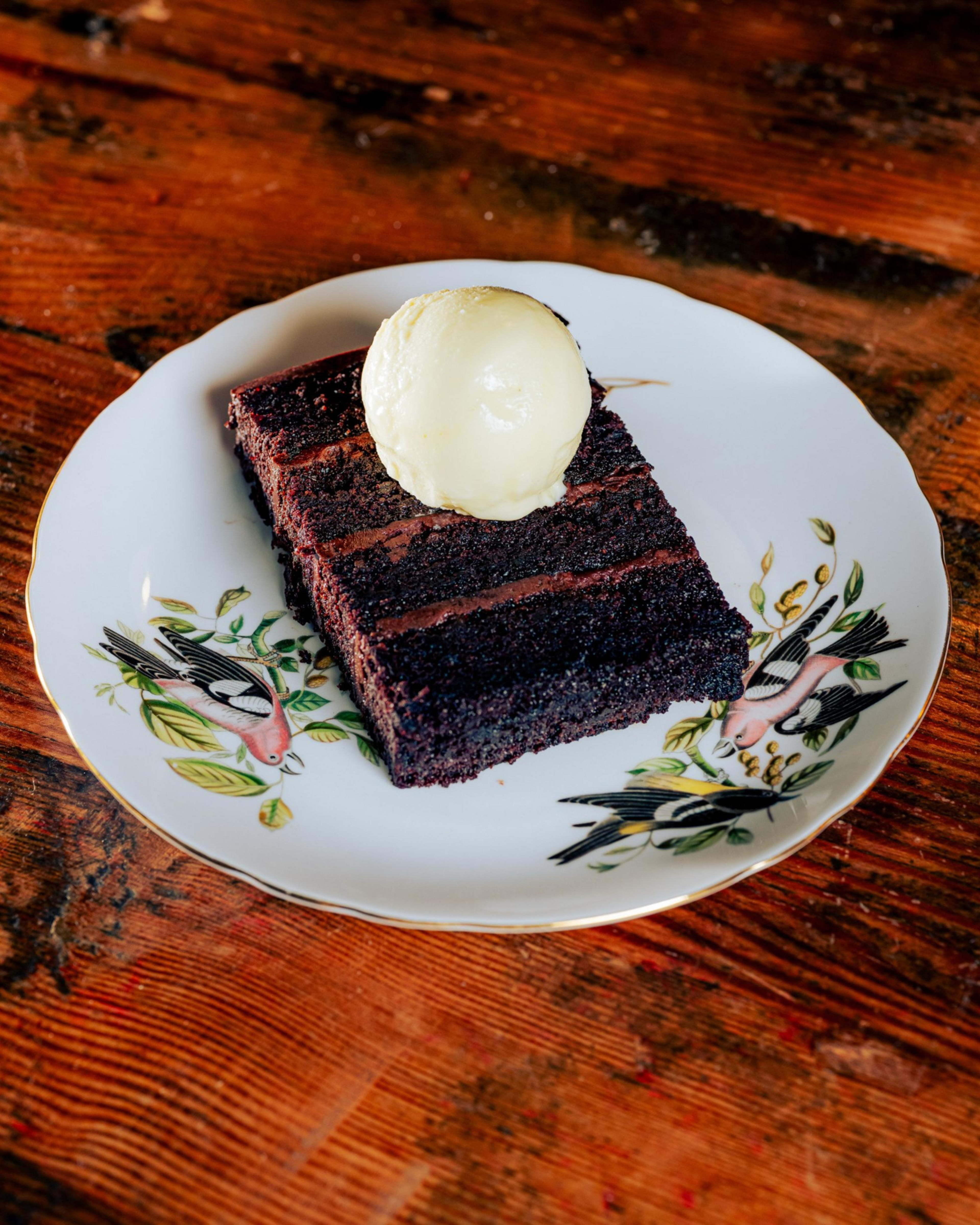 A slice of rich, dark chocolate cake with layers, topped with a scoop of vanilla ice cream is presented on a decorative plate featuring bird illustrations.