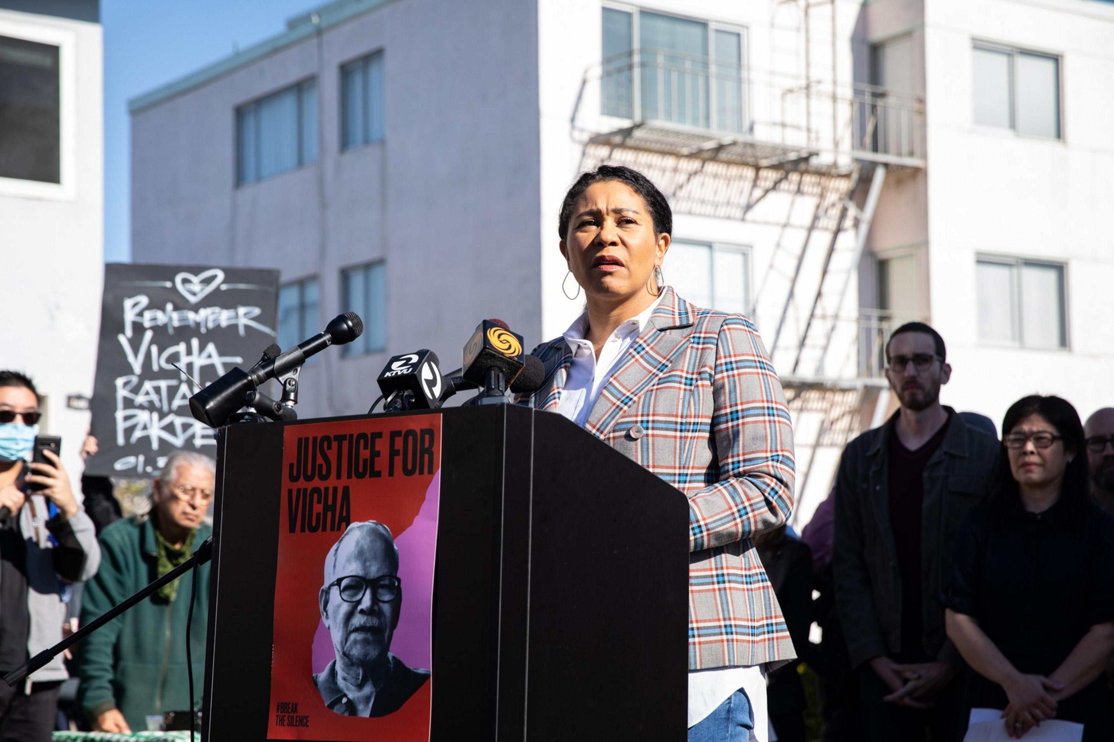 A woman speaks at a podium with microphones, featuring a poster that reads &quot;Justice for Vicha.&quot; A &quot;Remember Vicha Ratanapakdee&quot; sign is also visible.