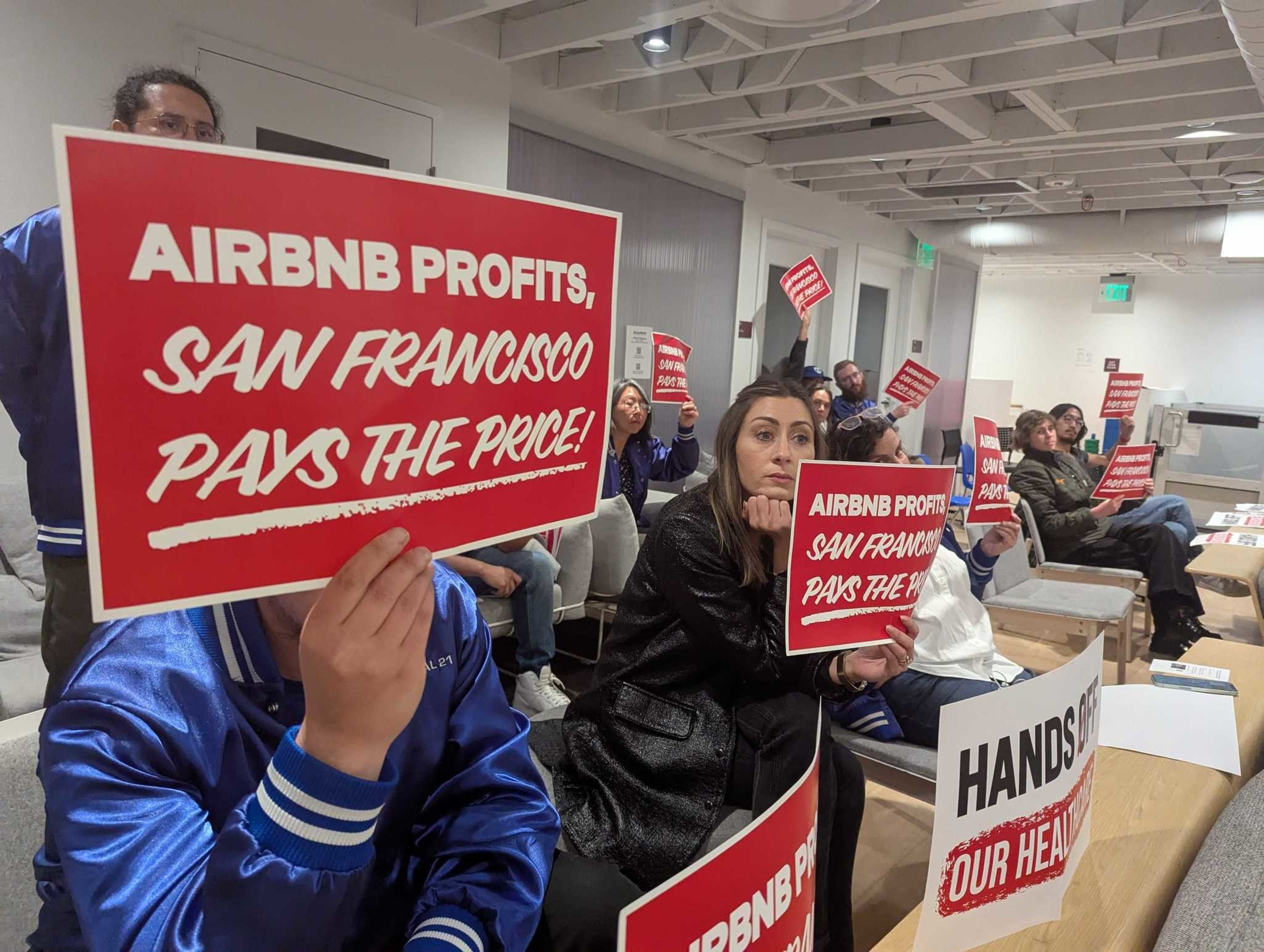 Workers in blue and black hold red protest signs reading &quot;Airbnb profits, San Francisco pays the price!&quot;