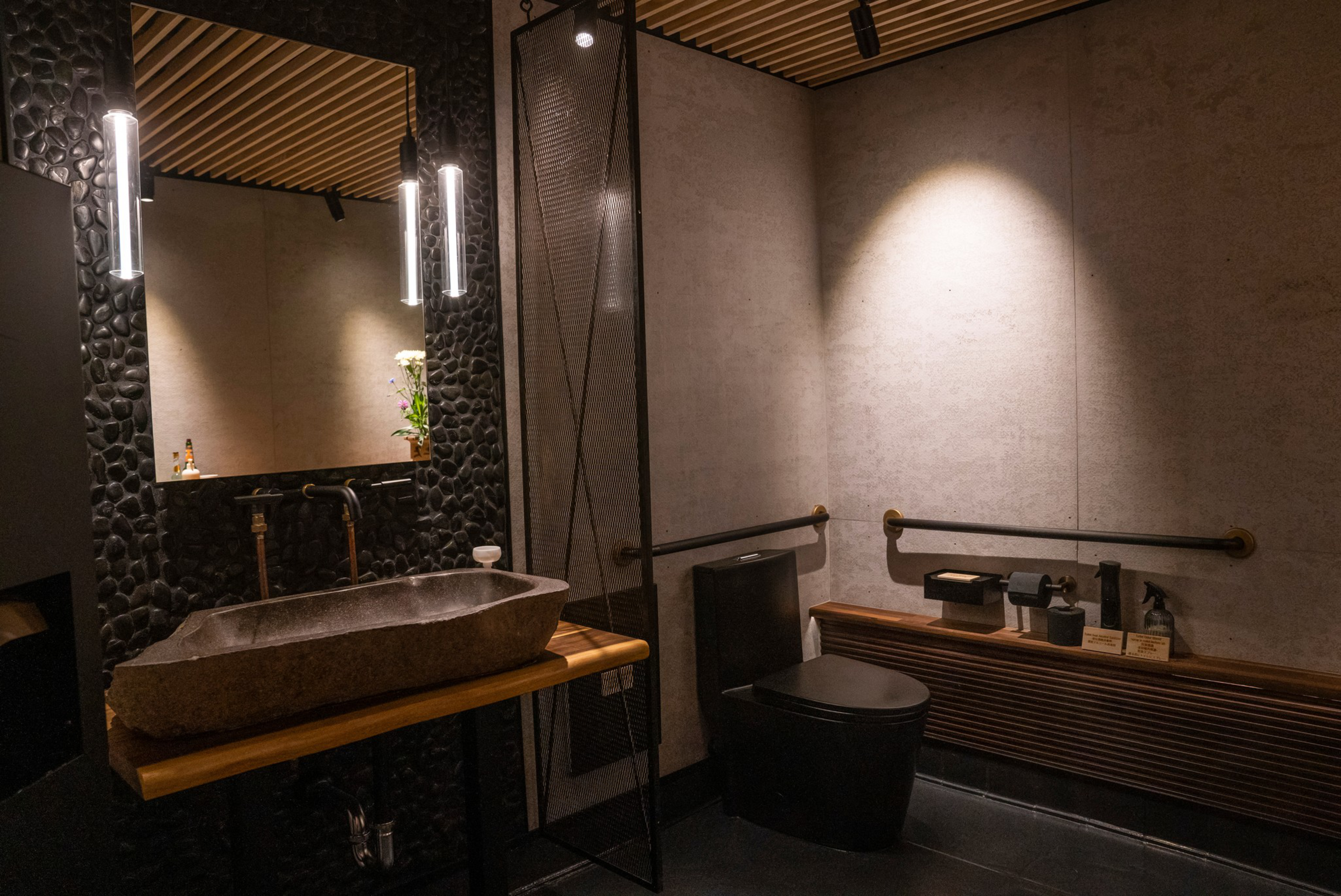 This image showcases a modern bathroom with a stone sink, textured walls, wooden accents, a large mirror, ambient lighting, and minimalistic black fixtures.