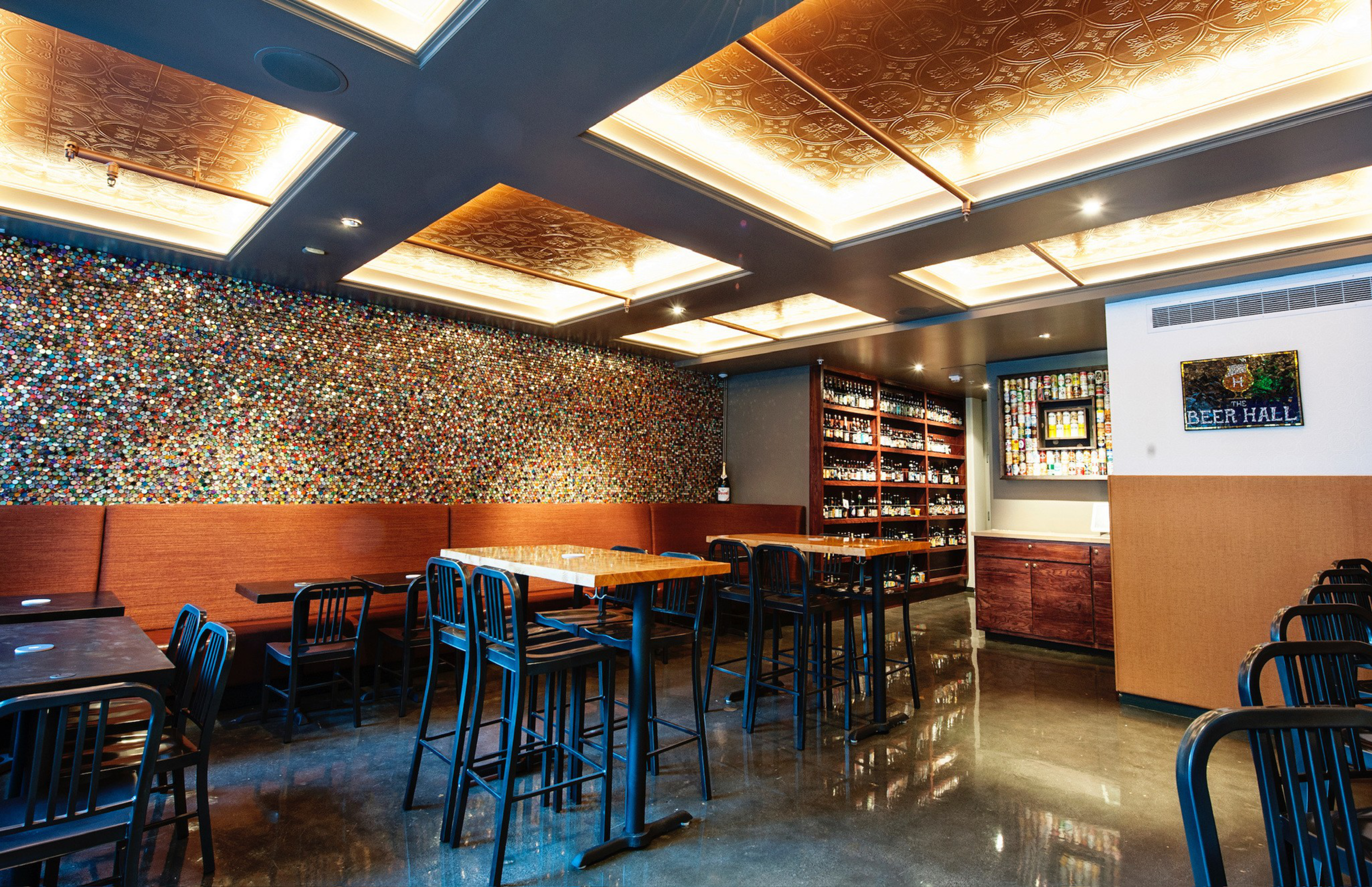 A cozy beer hall features wooden tables, black metal chairs, a mosaic bottle cap wall, shelves of liquor bottles, and warm ceiling lighting.