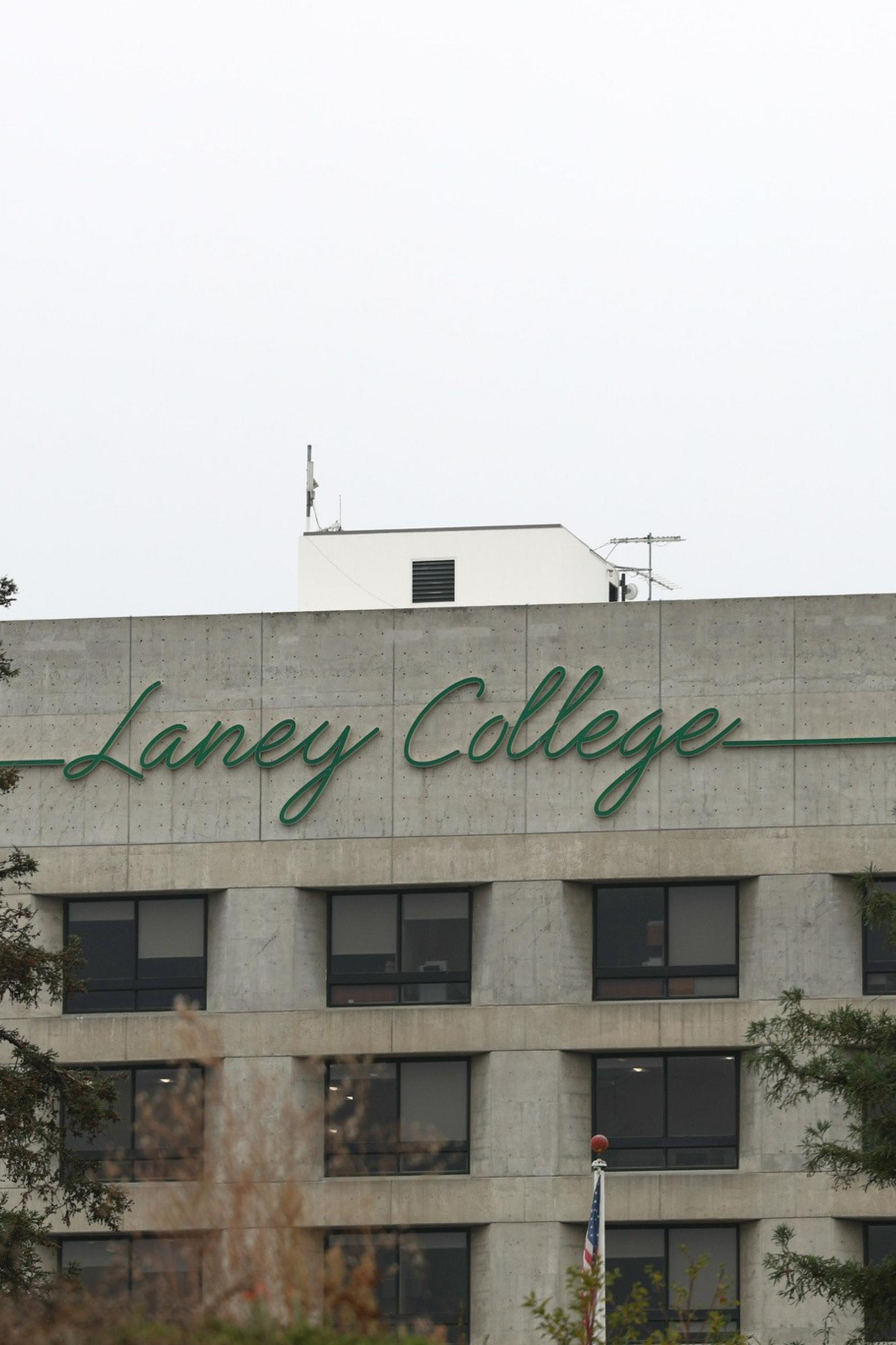A concrete building with "Laney College" written in green cursive lettering near the top, framed by tall evergreen trees on both sides.