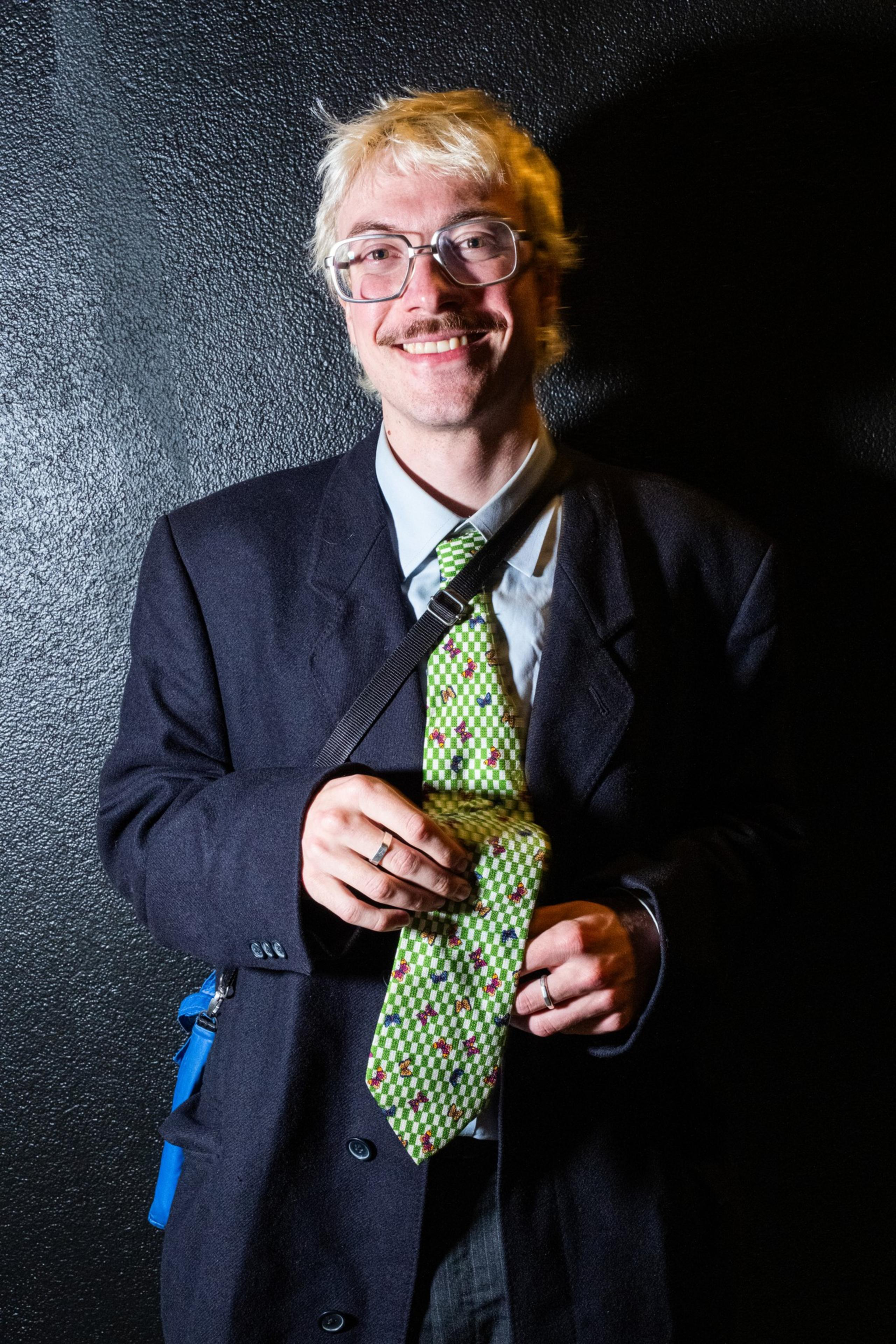 A person with light hair and glasses is smiling. They wear a dark blazer, a patterned green tie, and hold a strap over their shoulder against a dark backdrop.