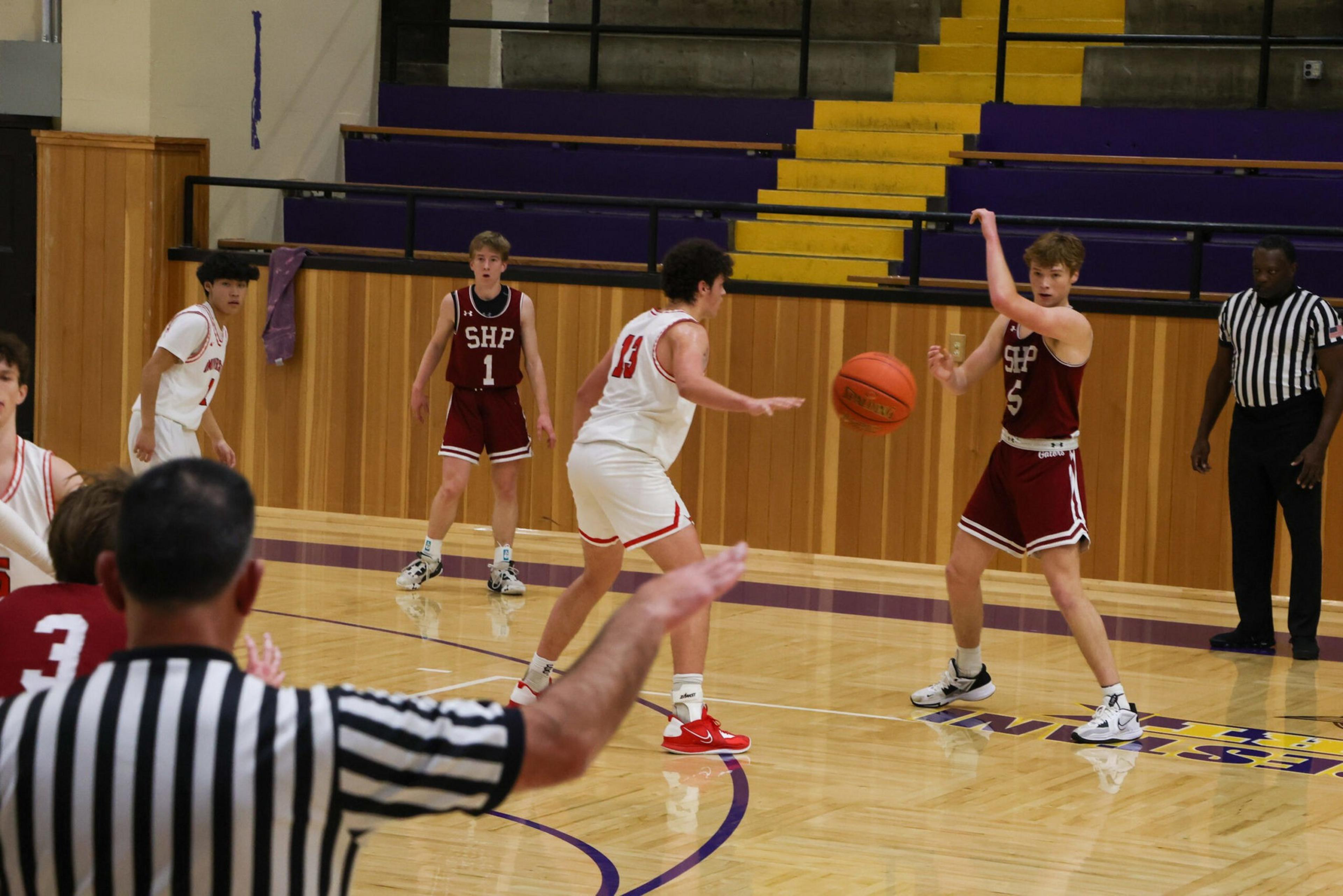 Two basketball teams compete on an indoor court, with players in white and maroon jerseys, and a referee signaling in the foreground.