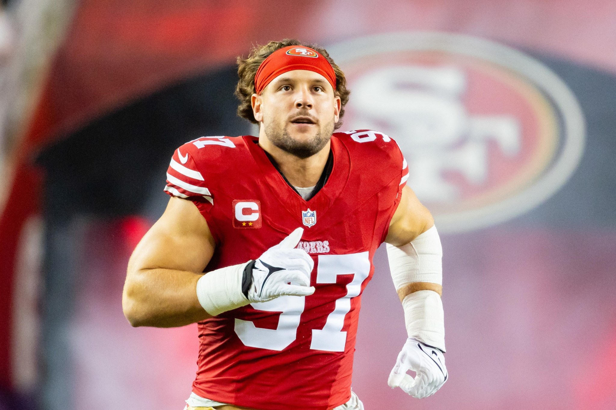 A football player in a red jersey with the number 97 is running on the field. He wears a red headband and has white gloves, with a large team logo in the background.