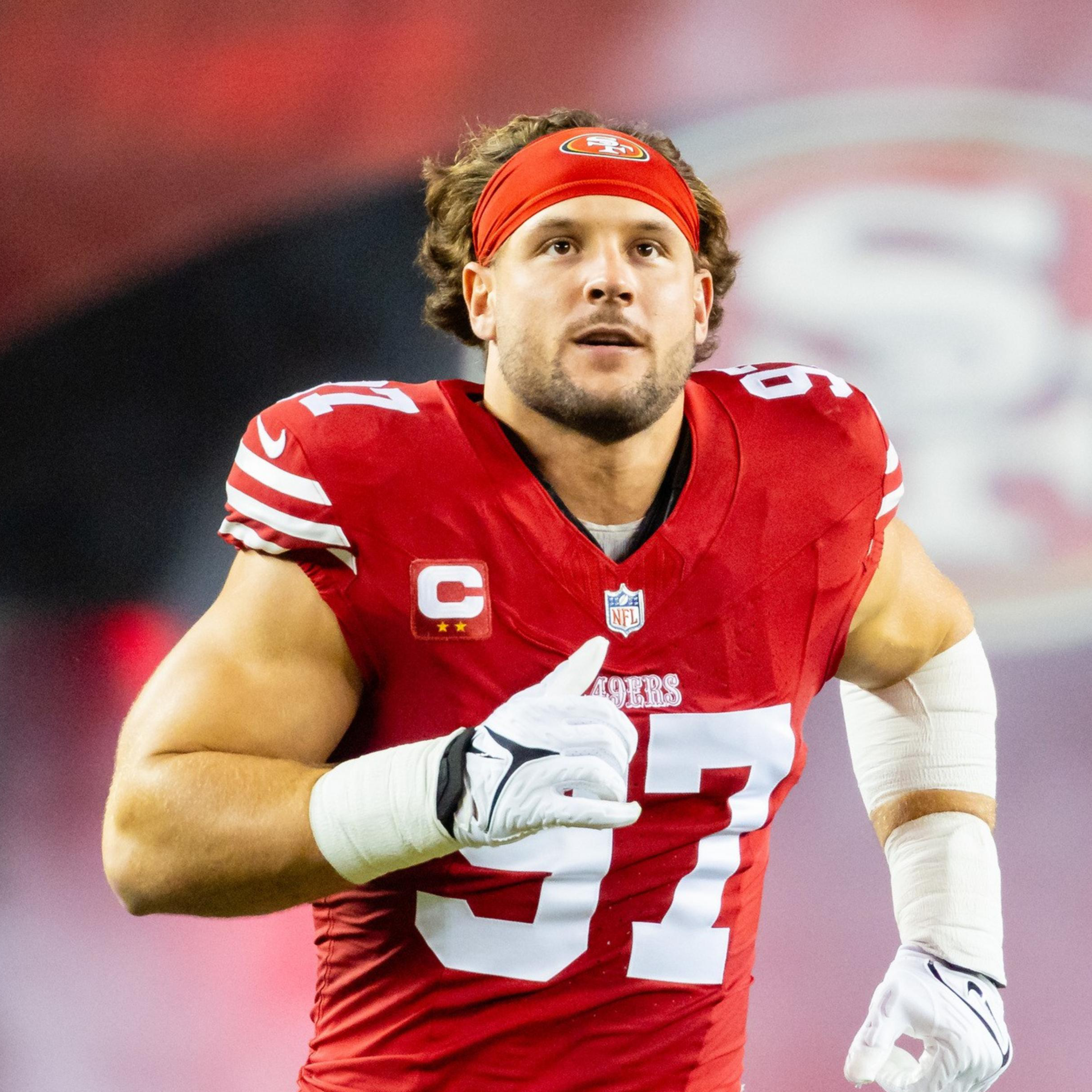 A football player in a red jersey with the number 97 is running on the field. He wears a red headband and has white gloves, with a large team logo in the background.