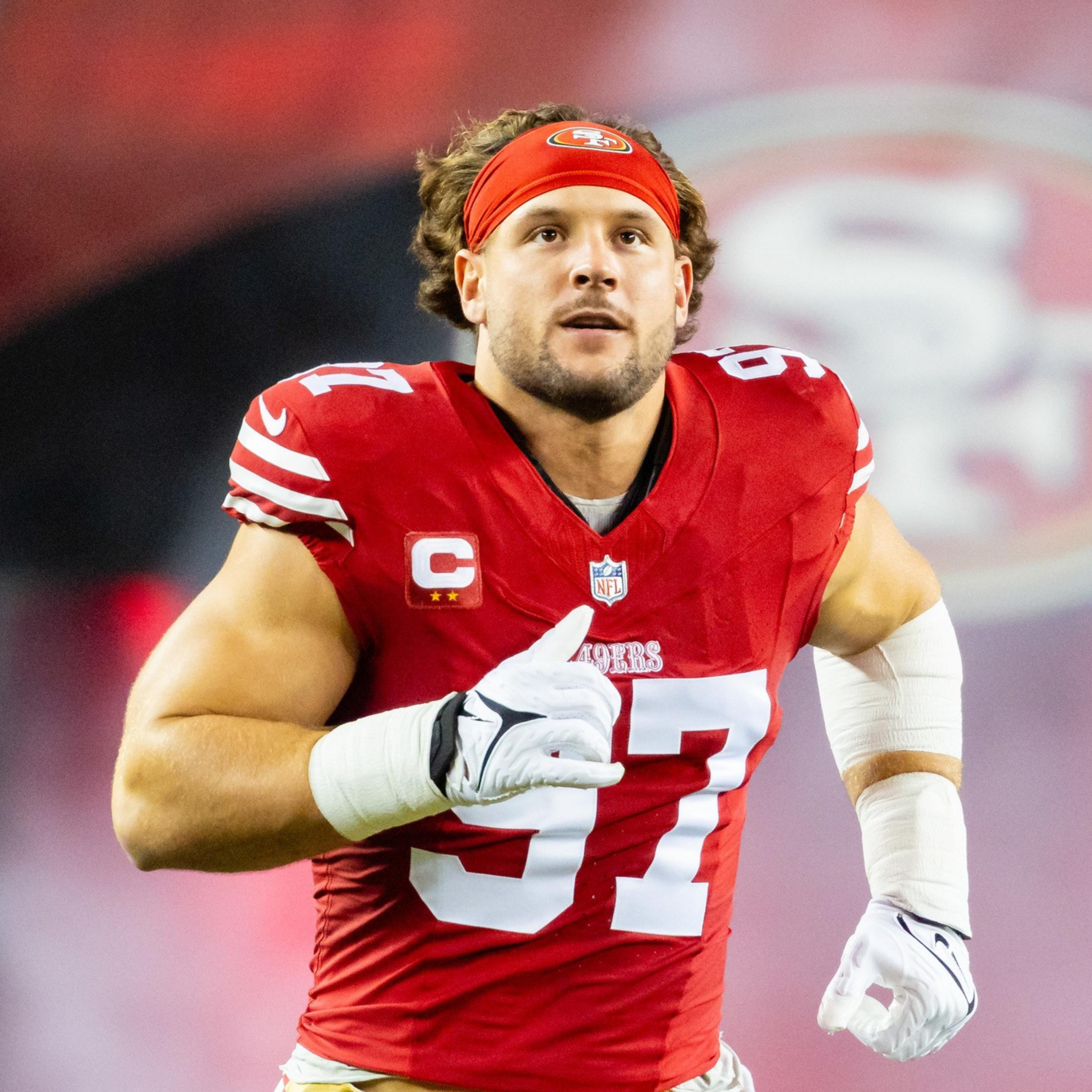 A football player in a red jersey with the number 97 is running on the field. He wears a red headband and has white gloves, with a large team logo in the background.