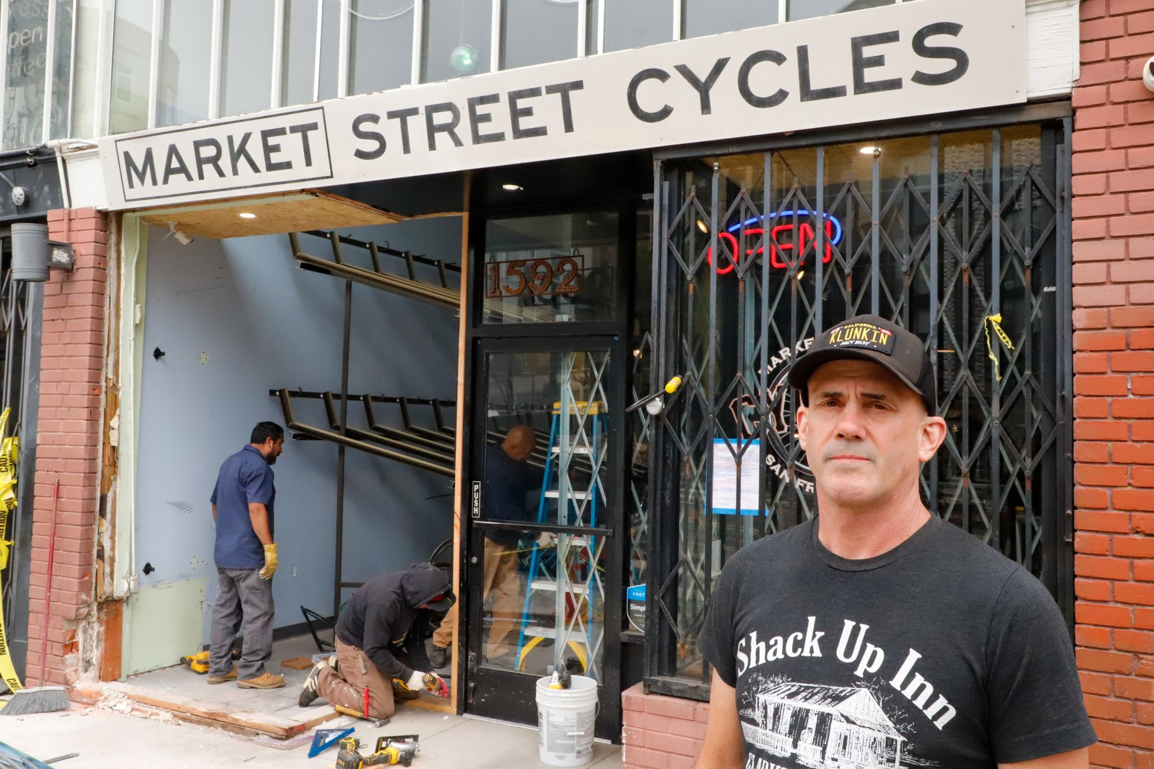 A man in a black shirt stands in front of Market Street Cycles. Two workers inside the shop, one standing and one kneeling, are renovating near an entrance. There's an "OPEN" sign in the window.