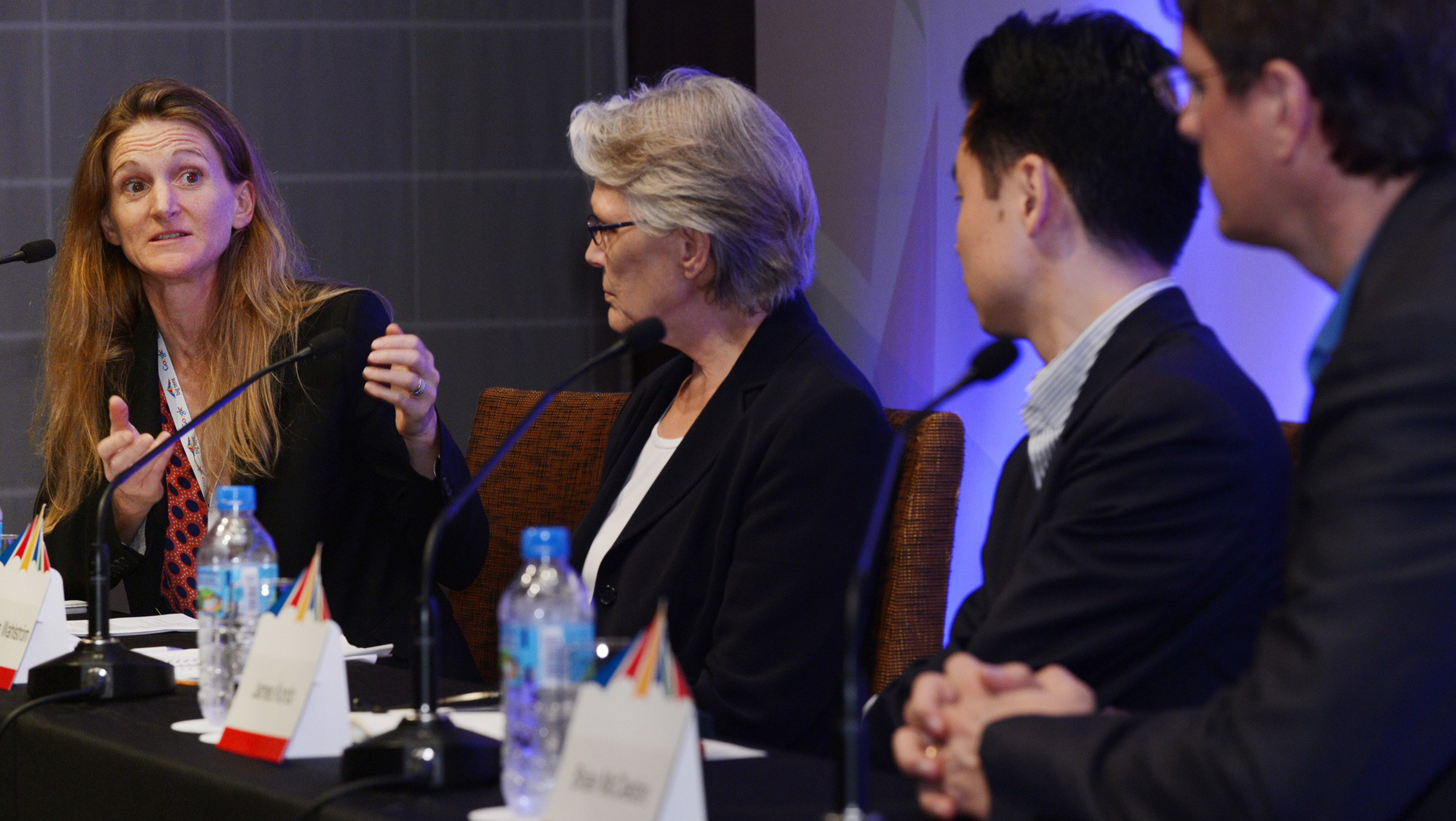 Four people sit at a panel table with microphones, water bottles, and name cards, engaged in a discussion at a formal event.