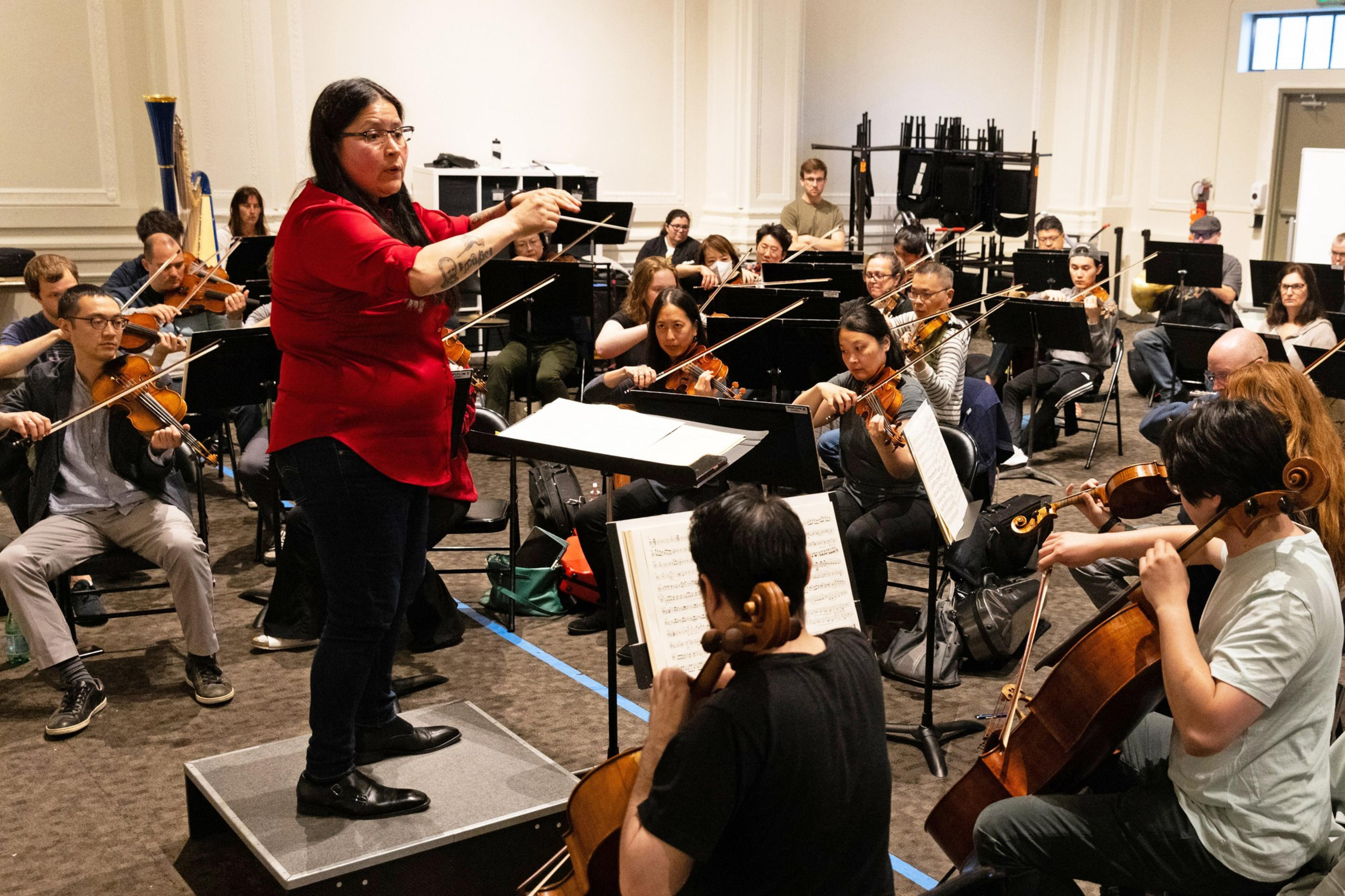 A conductor, wearing a red shirt, leads an orchestra in rehearsal, with musicians playing various string instruments and reading sheet music.