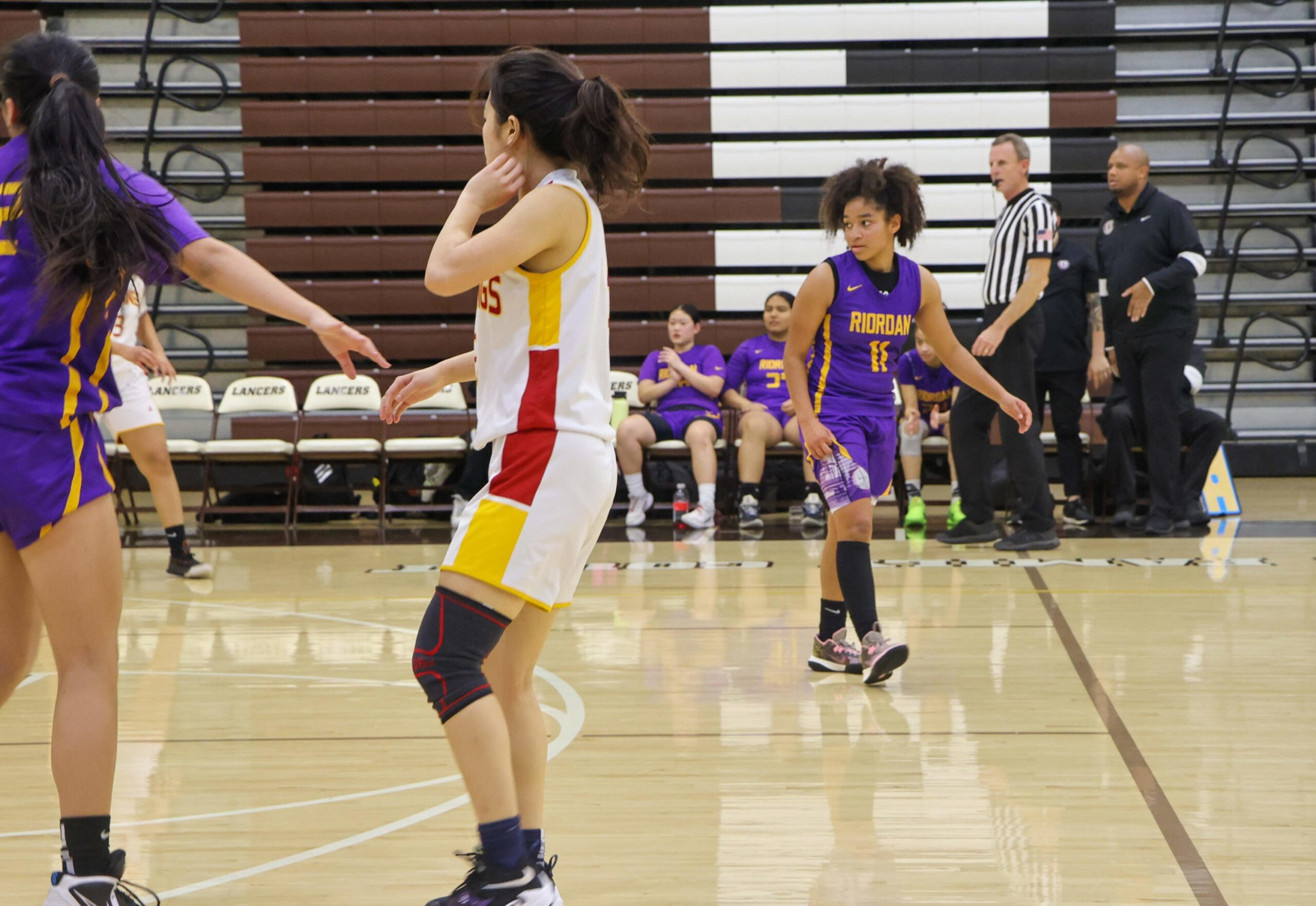 Two basketball players from opposing teams, one in white and yellow and the other in purple, prepare for play while a referee and coach stand near the sidelines.