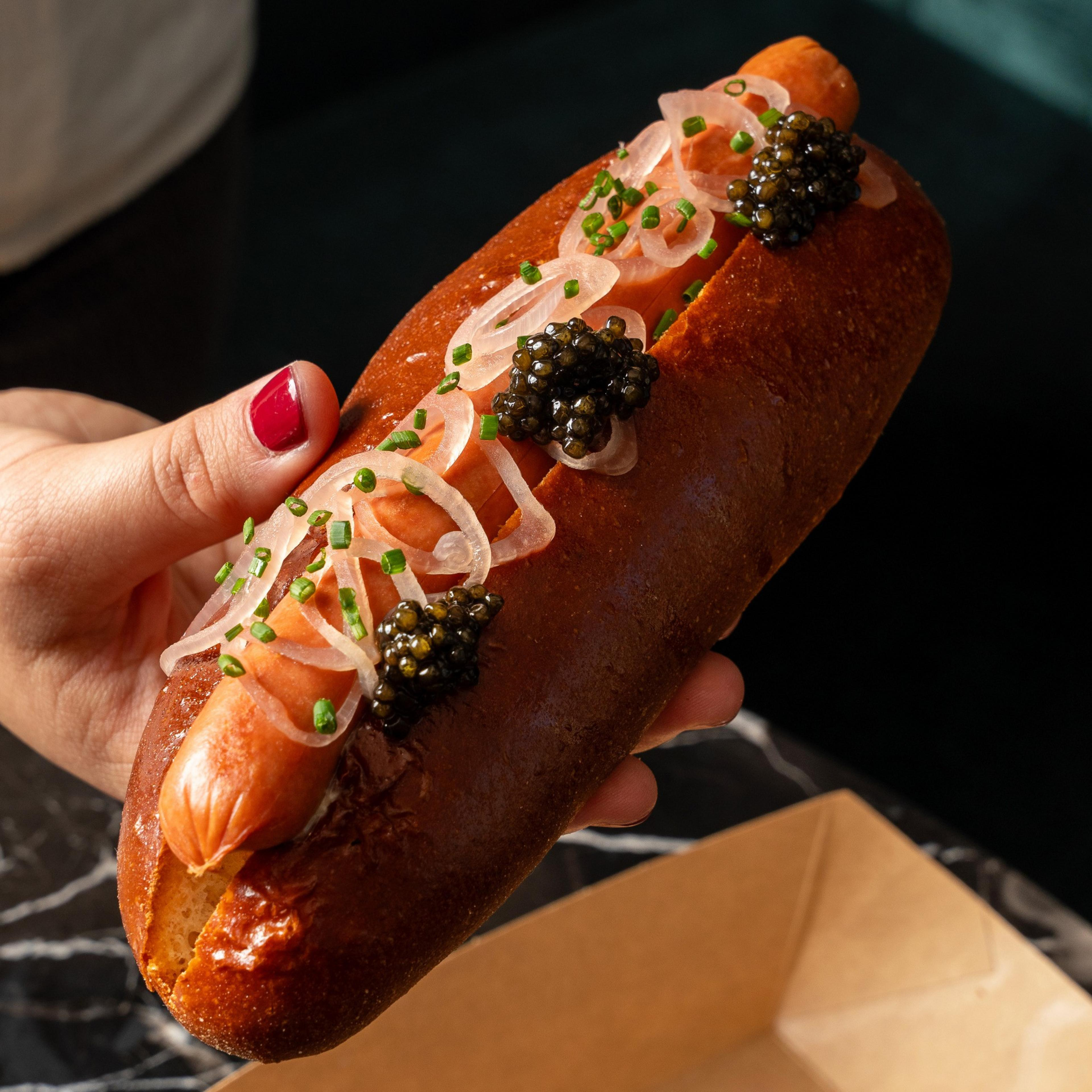A person holds a gourmet hot dog in a shiny bun, topped with caviar, thin onion slices, and chopped chives, with a box in the background.