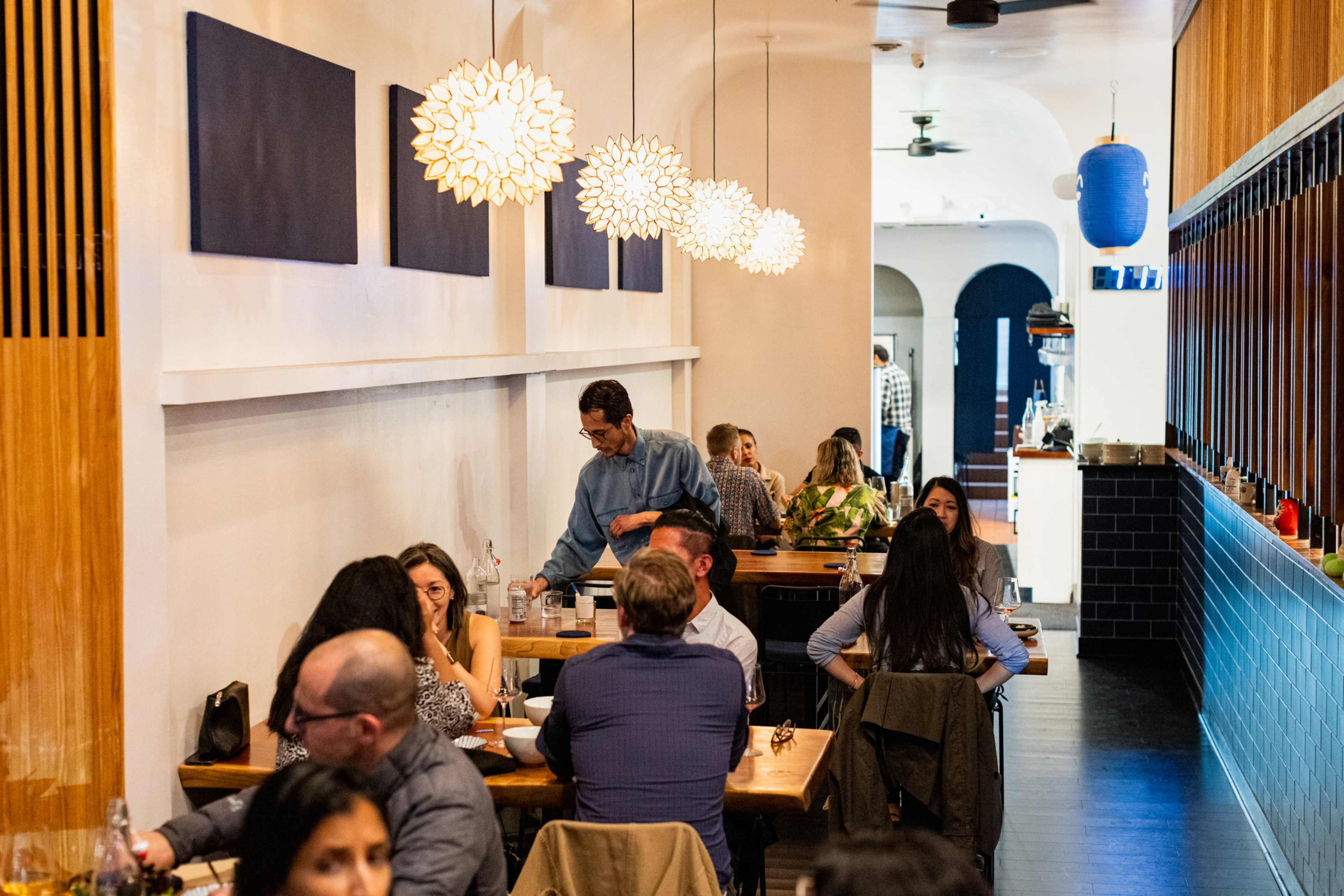 People are dining at wooden tables in a modern restaurant with pendant floral lights, white walls, blue tile accents, and a server attending to guests.