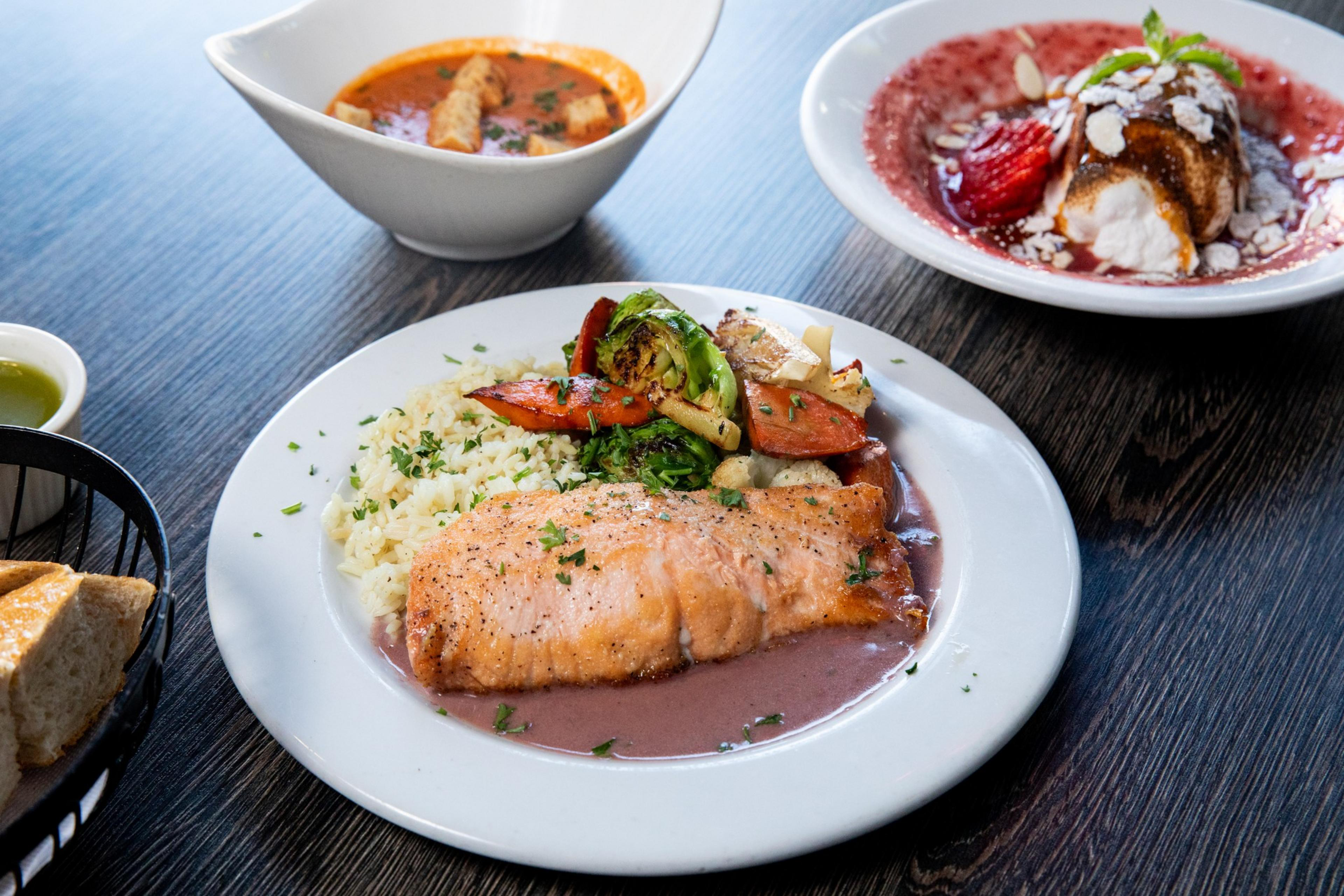 The image shows a salmon fillet with rice and vegetables on a white plate, a bowl of soup, a dessert with strawberries and cream, and a basket of bread on a dark wooden table.