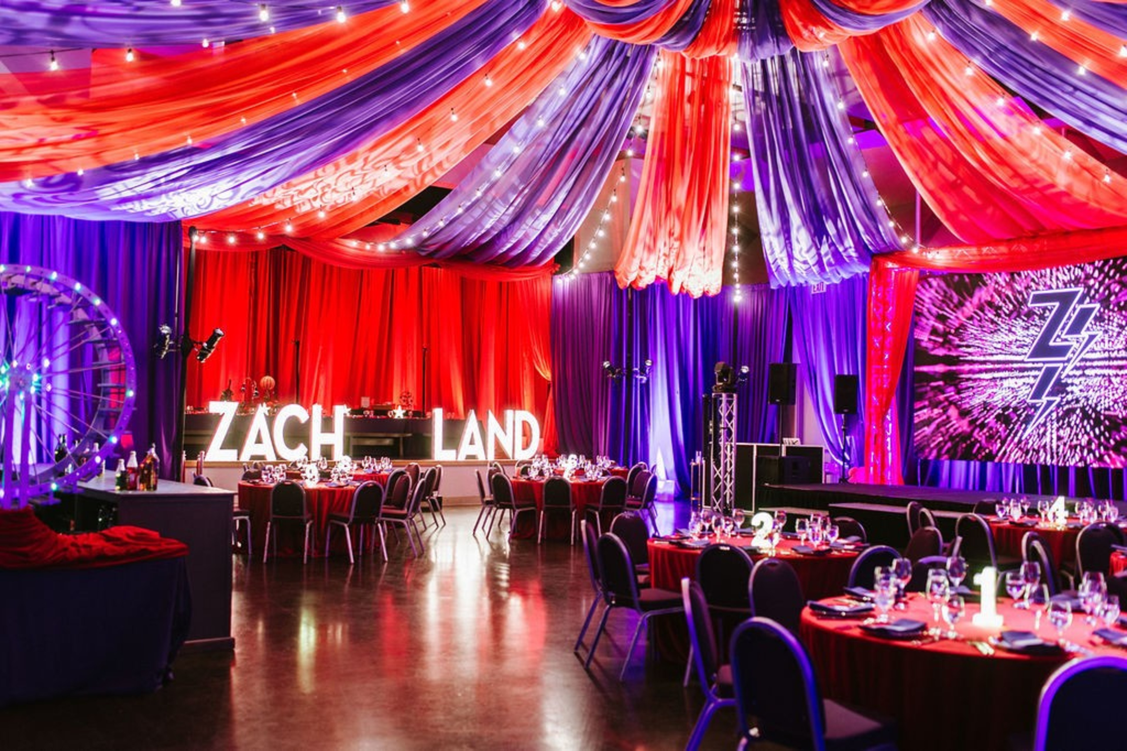 The image shows a vibrant event space adorned with red and purple drapes, glowing â€œZACH LANDâ€ letters, round tables with elegant settings, and a colorful stage backdrop.