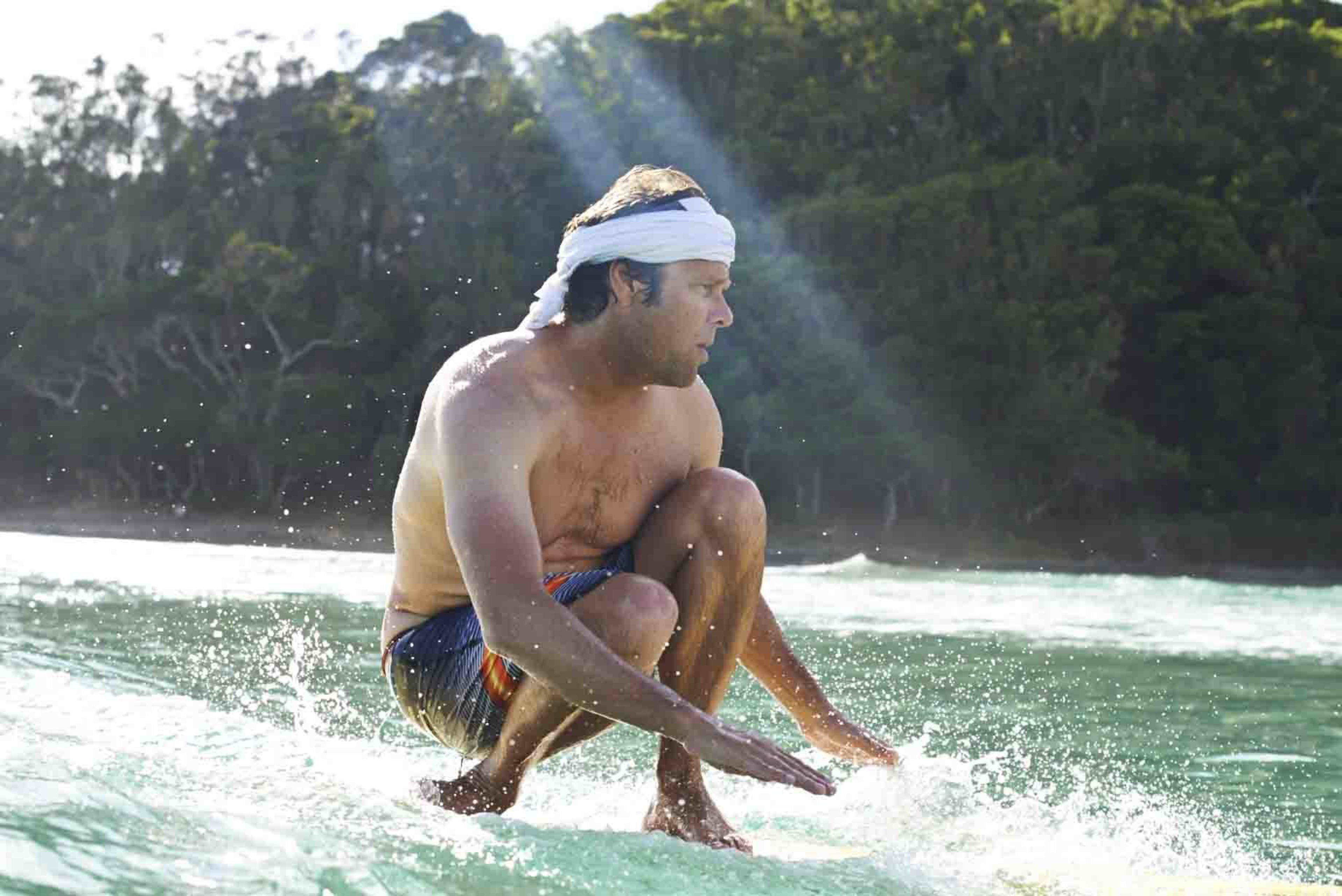 A shirtless man wearing colorful shorts and a white headband crouches low while surfing on a wave near a forested shoreline.