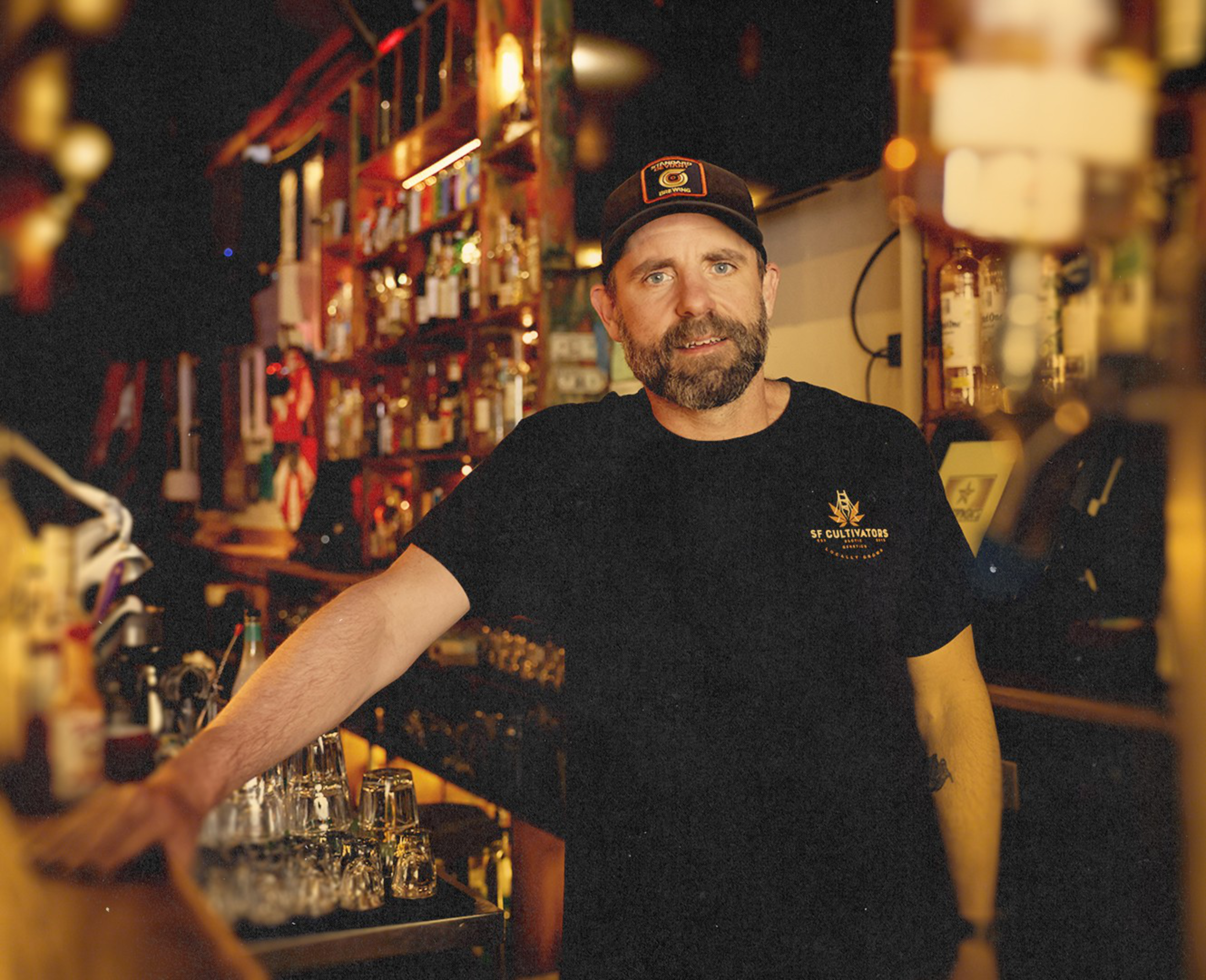 A bearded man wearing a black t-shirt and cap stands behind a bar with shelves of liquor bottles in warm lighting.