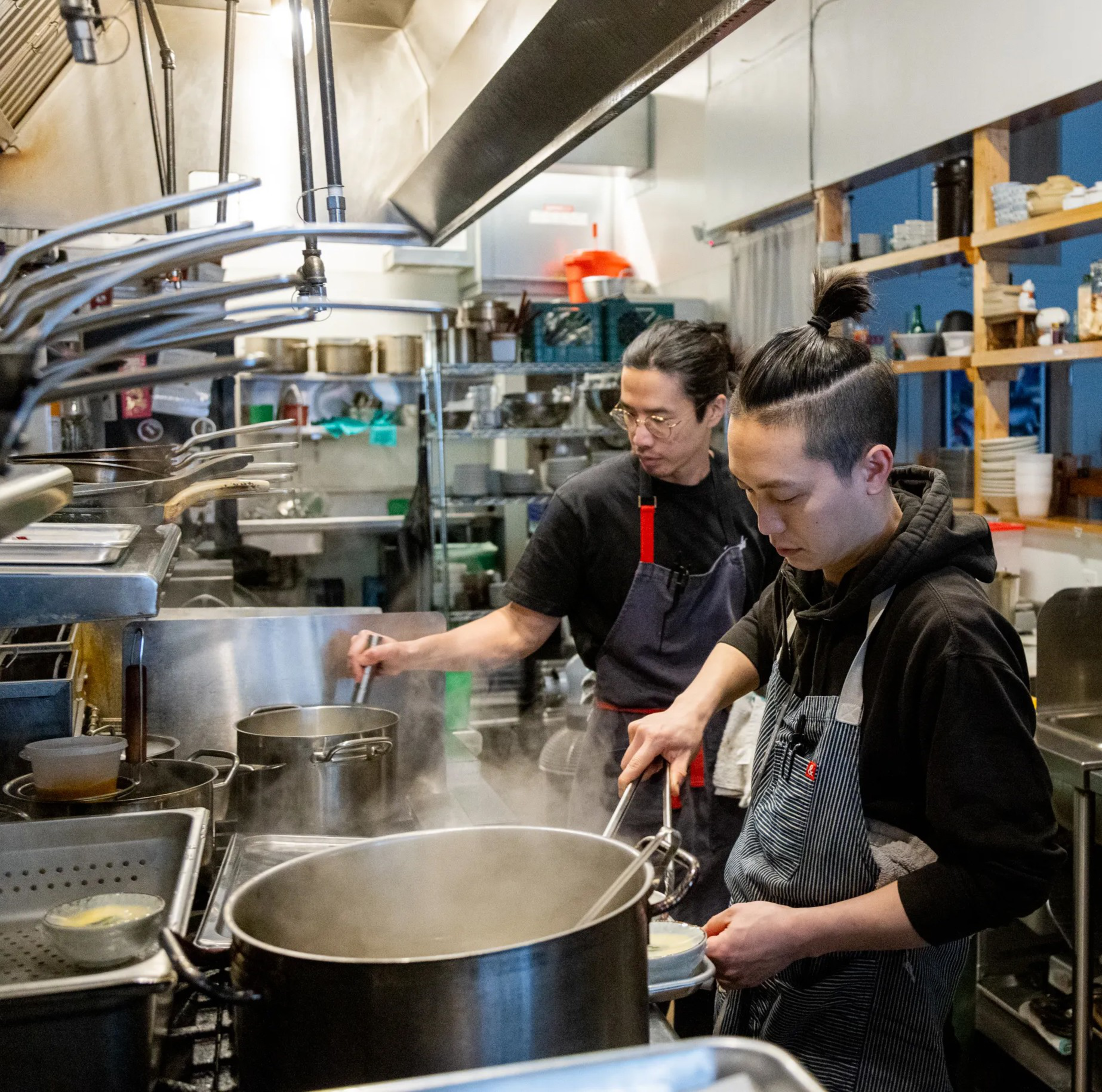 Two chefs are working in a busy commercial kitchen with pots steaming on the stove.