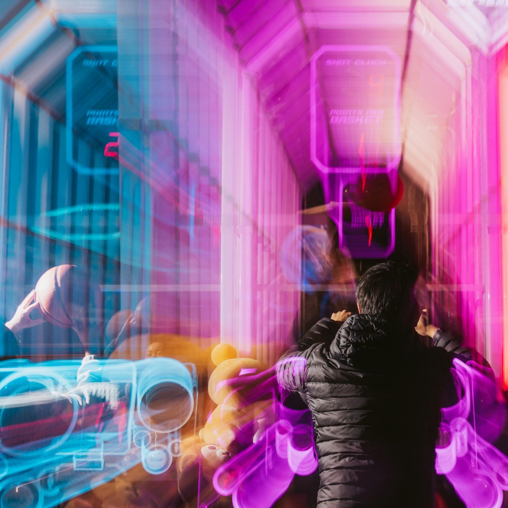 The image shows a person in a dark jacket standing in a vibrant, futuristic arcade with bright pink and blue neon lights, creating a dynamic and colorful blur effect.