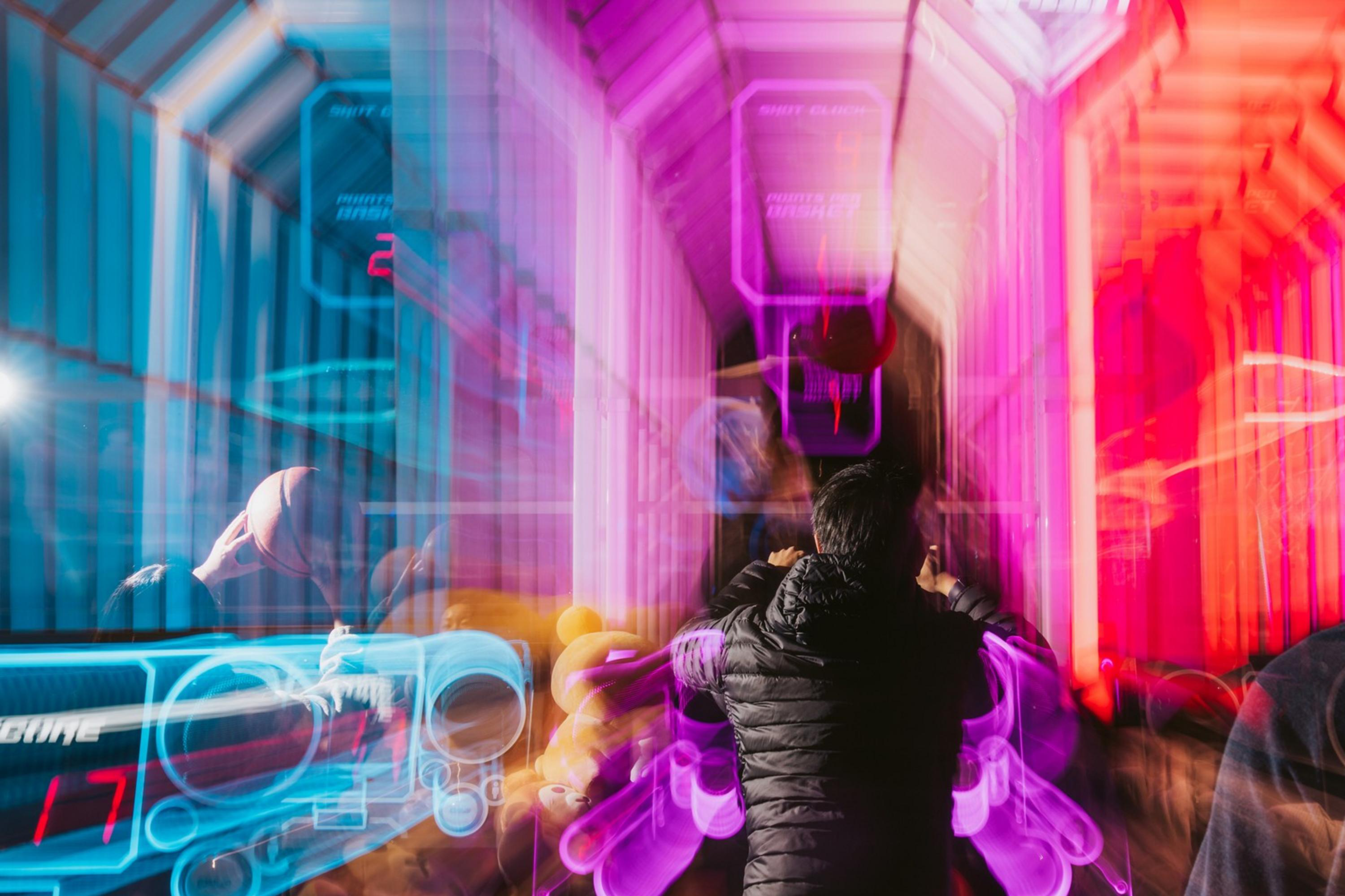 The image shows a person in a dark jacket standing in a vibrant, futuristic arcade with bright pink and blue neon lights, creating a dynamic and colorful blur effect.