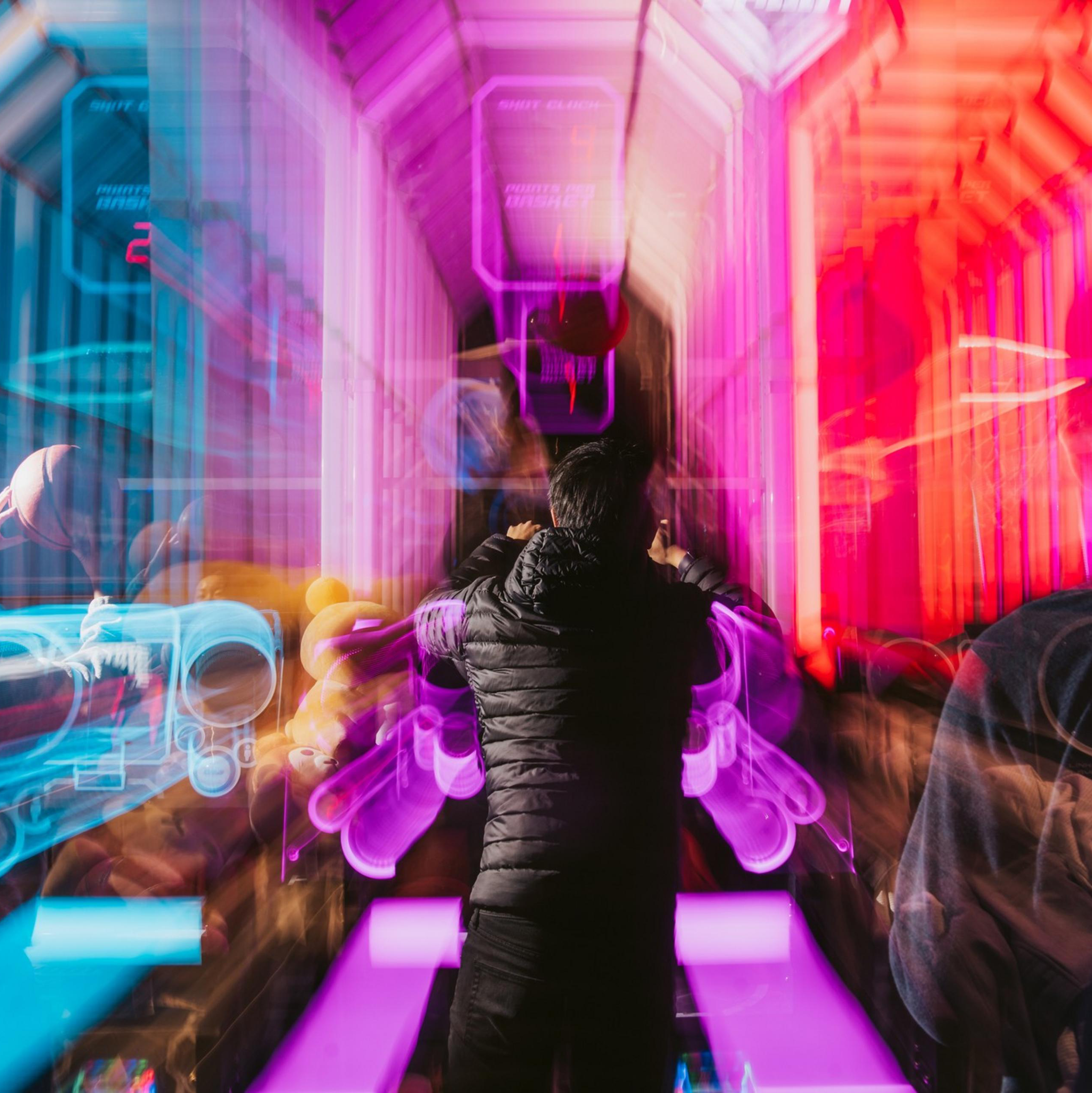 The image shows a person in a dark jacket standing in a vibrant, futuristic arcade with bright pink and blue neon lights, creating a dynamic and colorful blur effect.