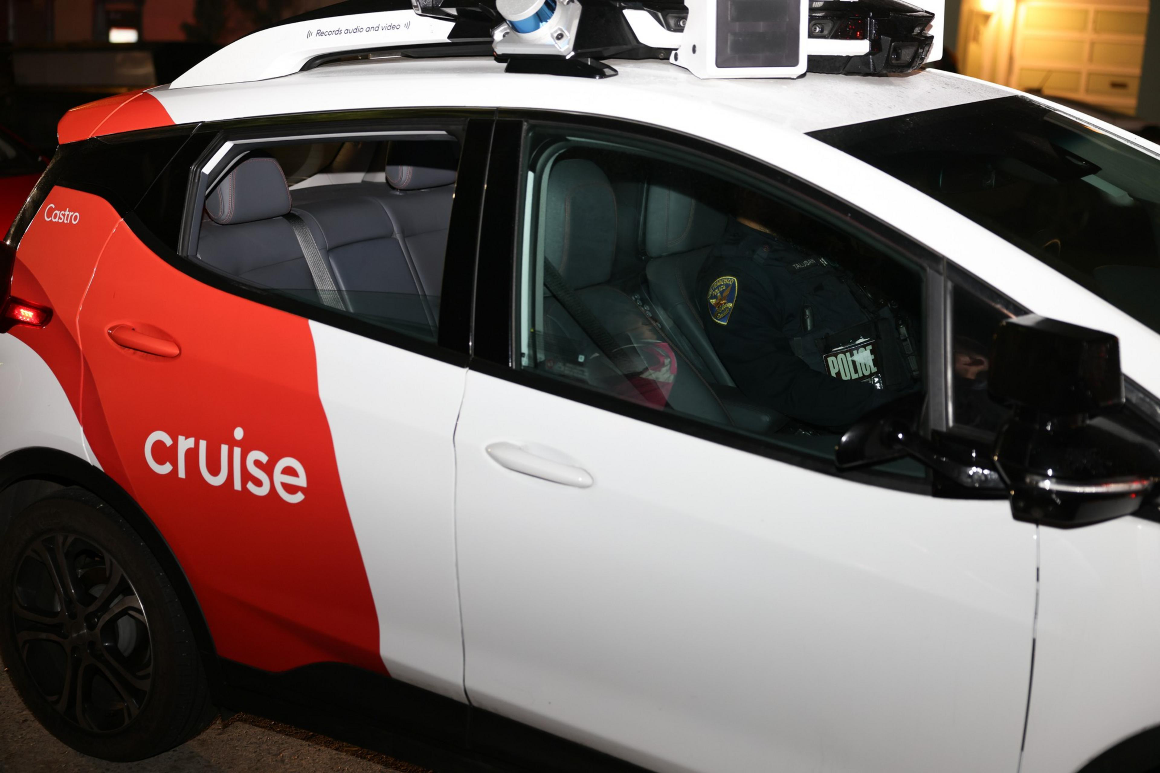 A white and red Cruise self-driving car is parked, with sensors on the roof and a police officer visible inside at the driver's seat.