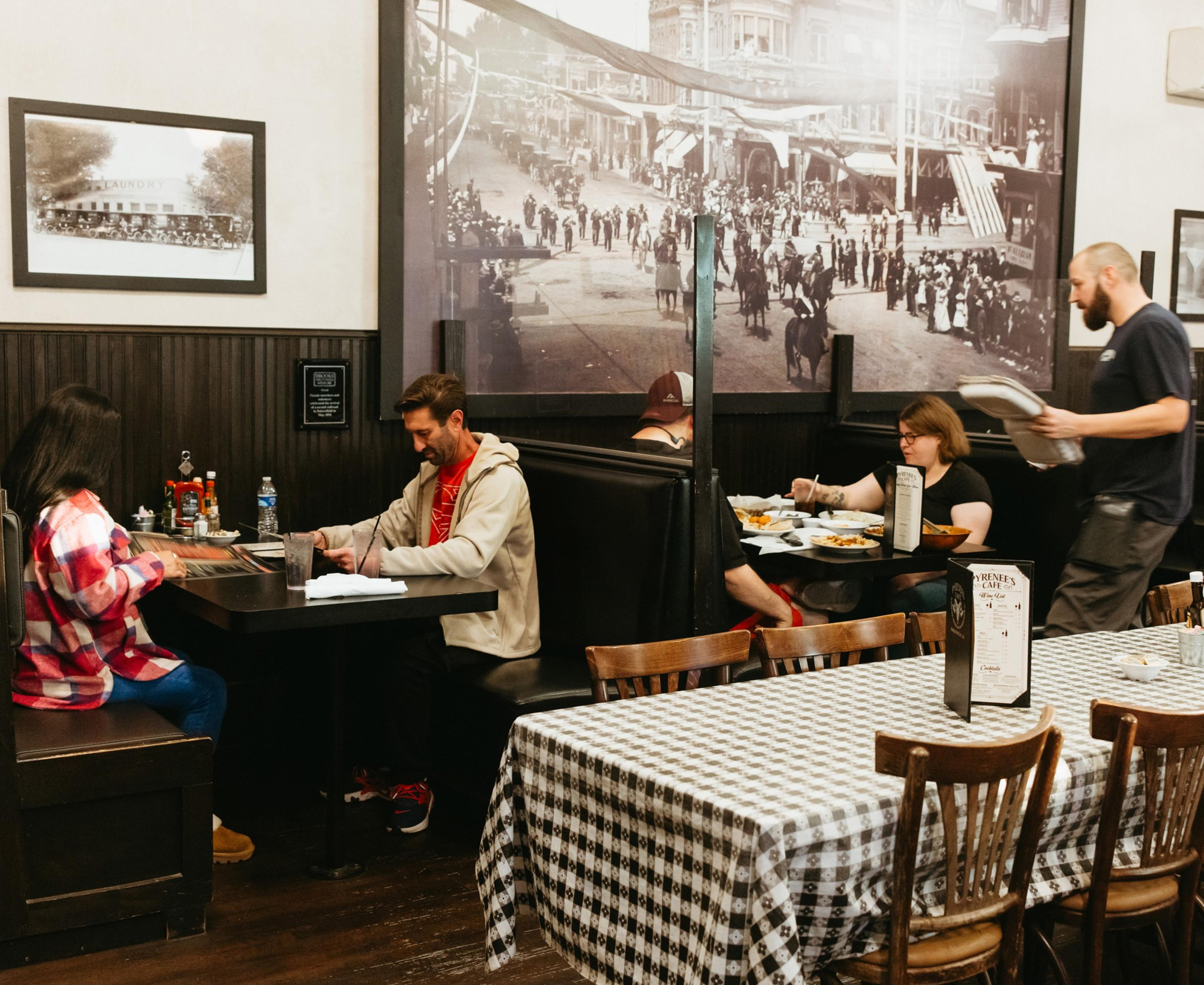 A cozy restaurant features people dining in booths, with a black-and-white historic photo on the wall. A server is delivering food, and a checkered tablecloth is seen.
