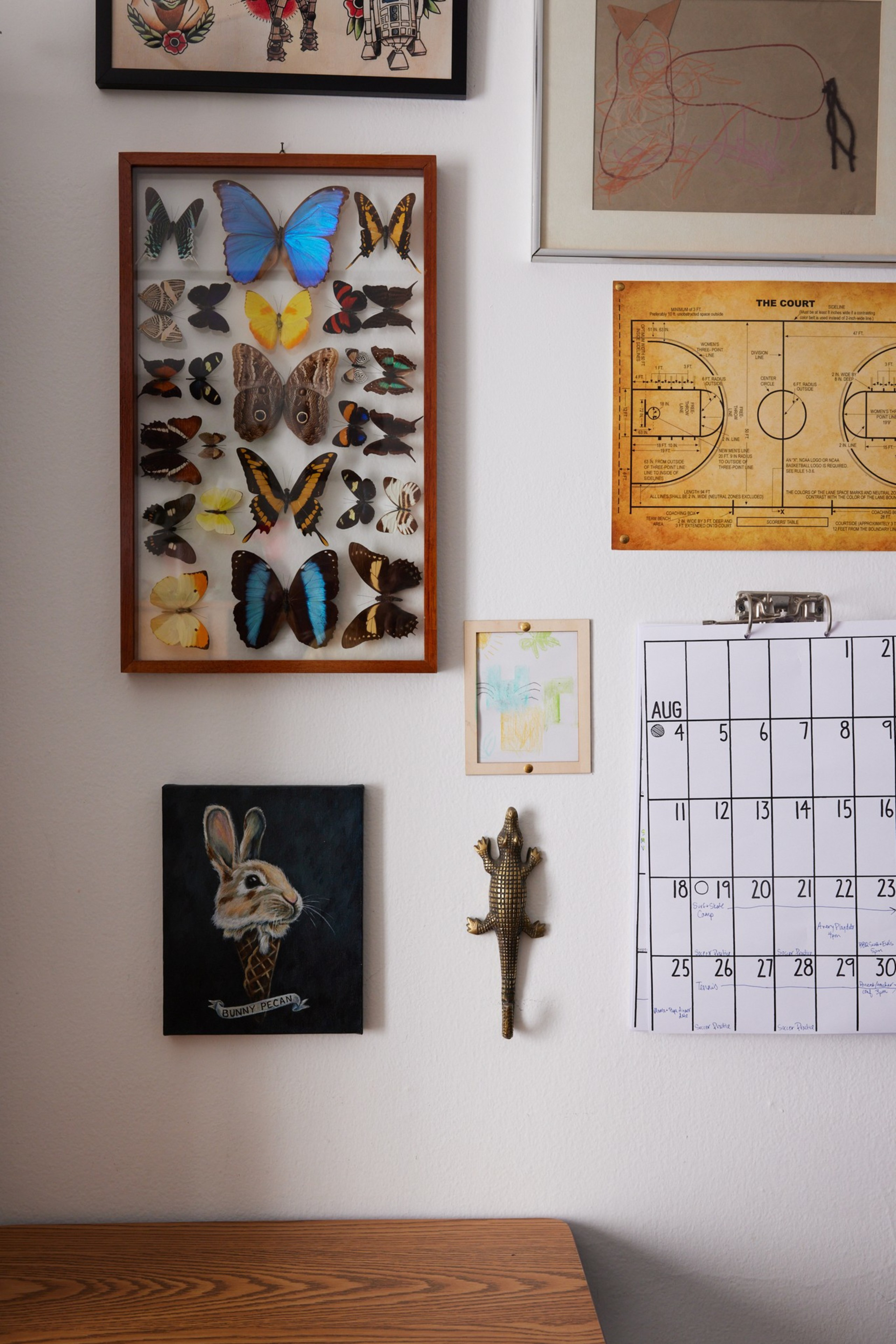 A wall features framed butterflies, art, a lizard sculpture, a calendar, and a drawing; below is a wooden desk.