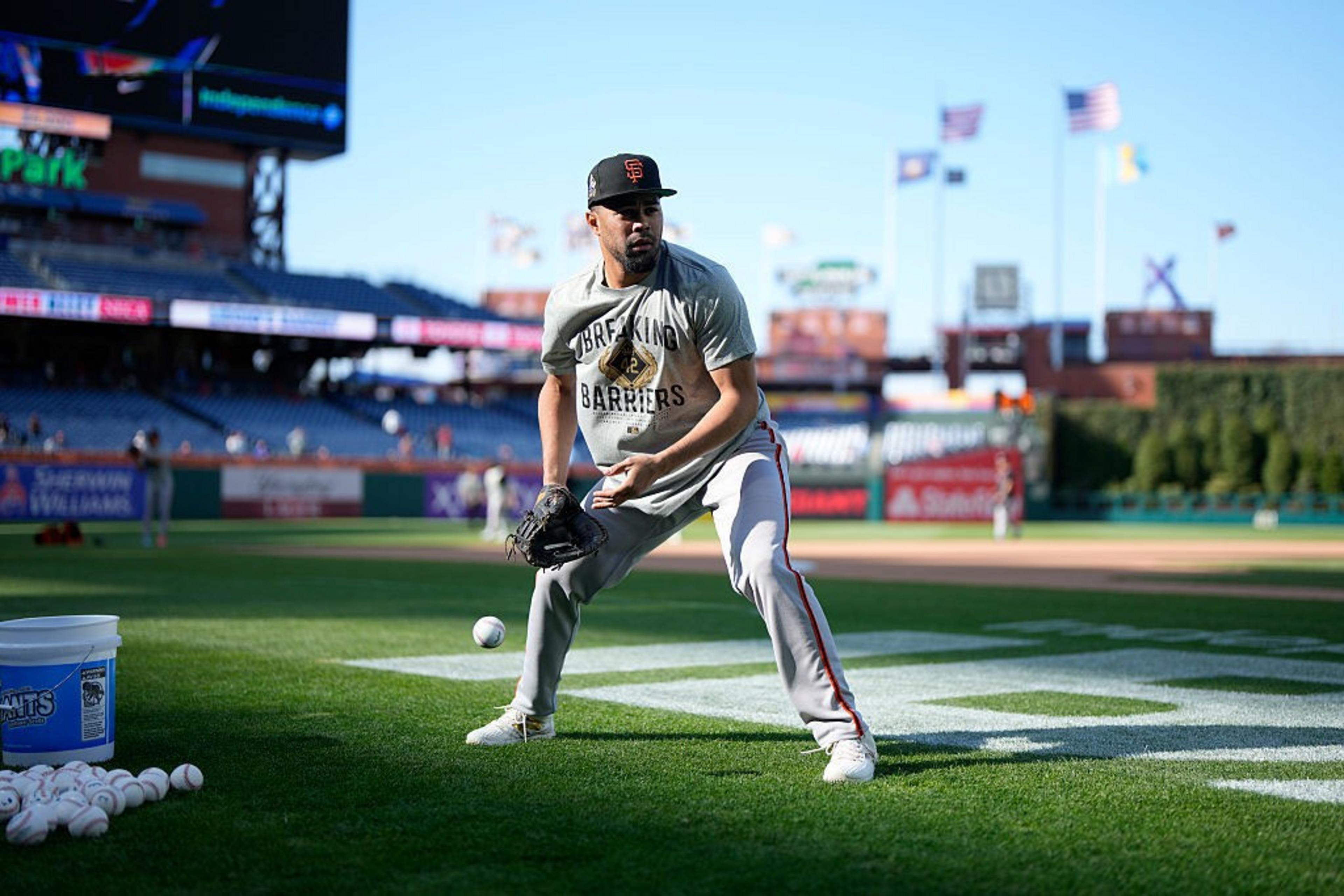 A baseball player in a &quot;Breaking Barriers&quot; shirt practices fielding on a grassy field. A bucket of baseballs sits nearby. The sky is clear, with flags waving.