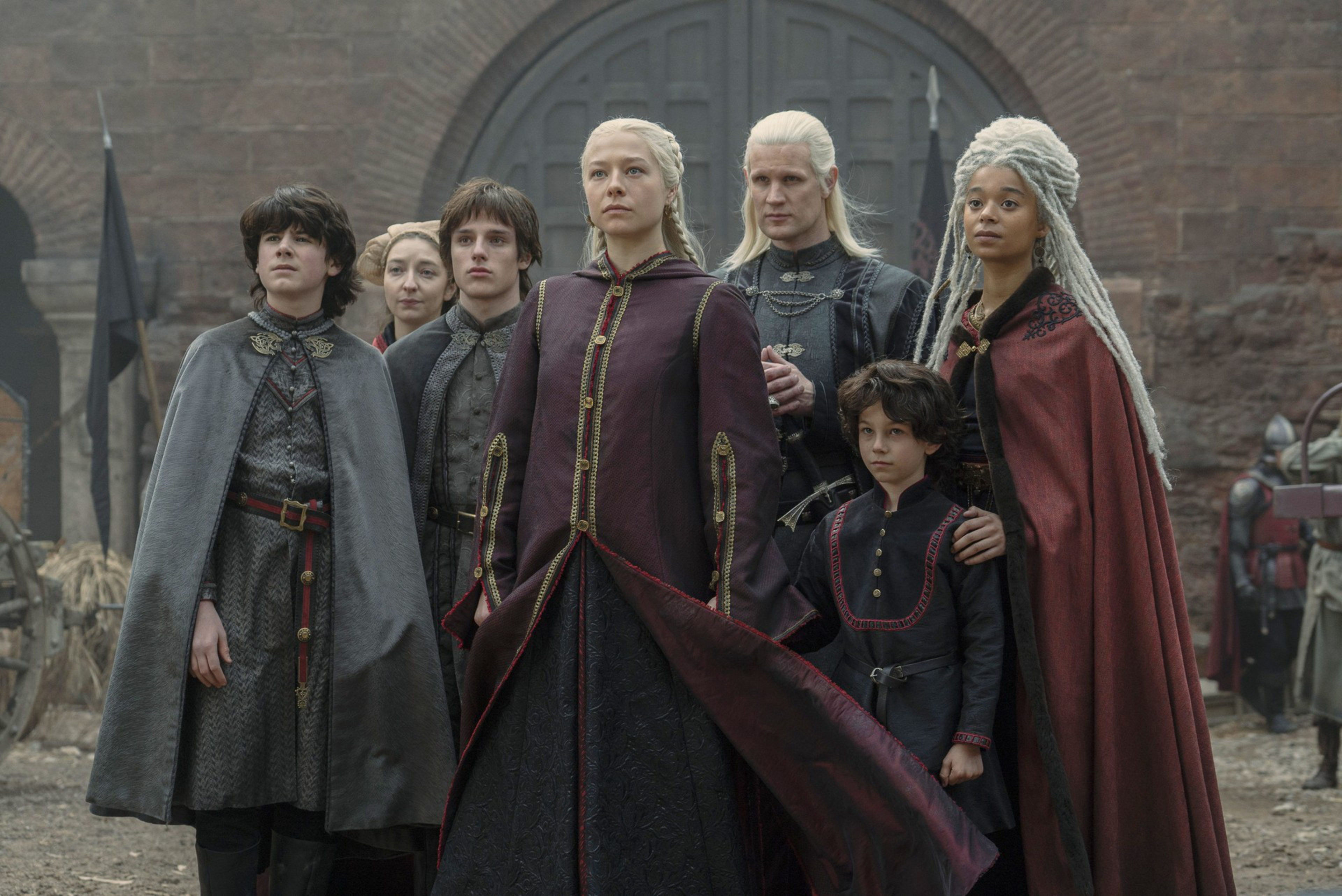 Seven young people dressed in medieval-style robes and cloaks stand together in front of a large stone building with a wooden door.