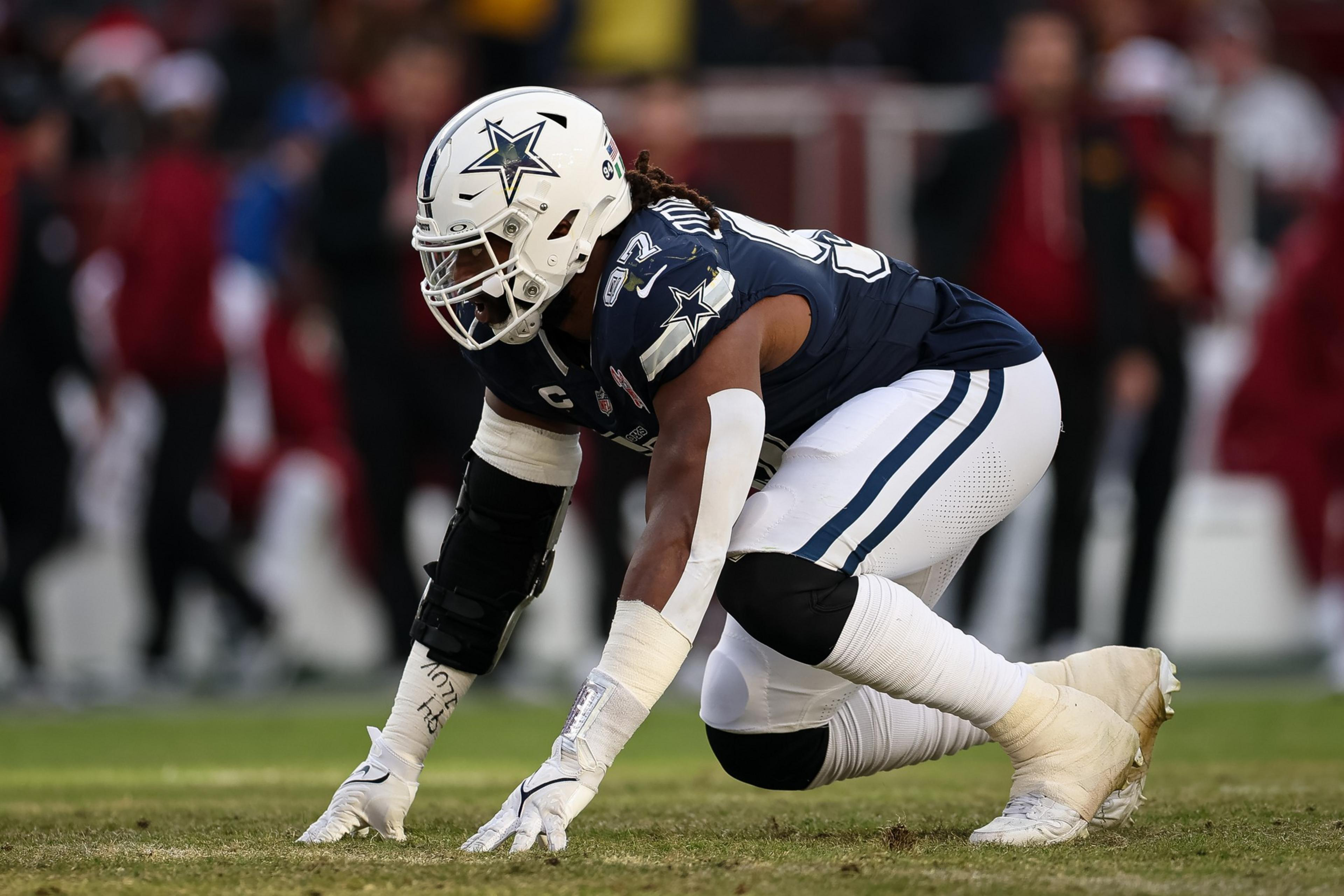 A Dallas Cowboys football player in full gear crouches on the field, ready to start a play, with blurred spectators in the background.