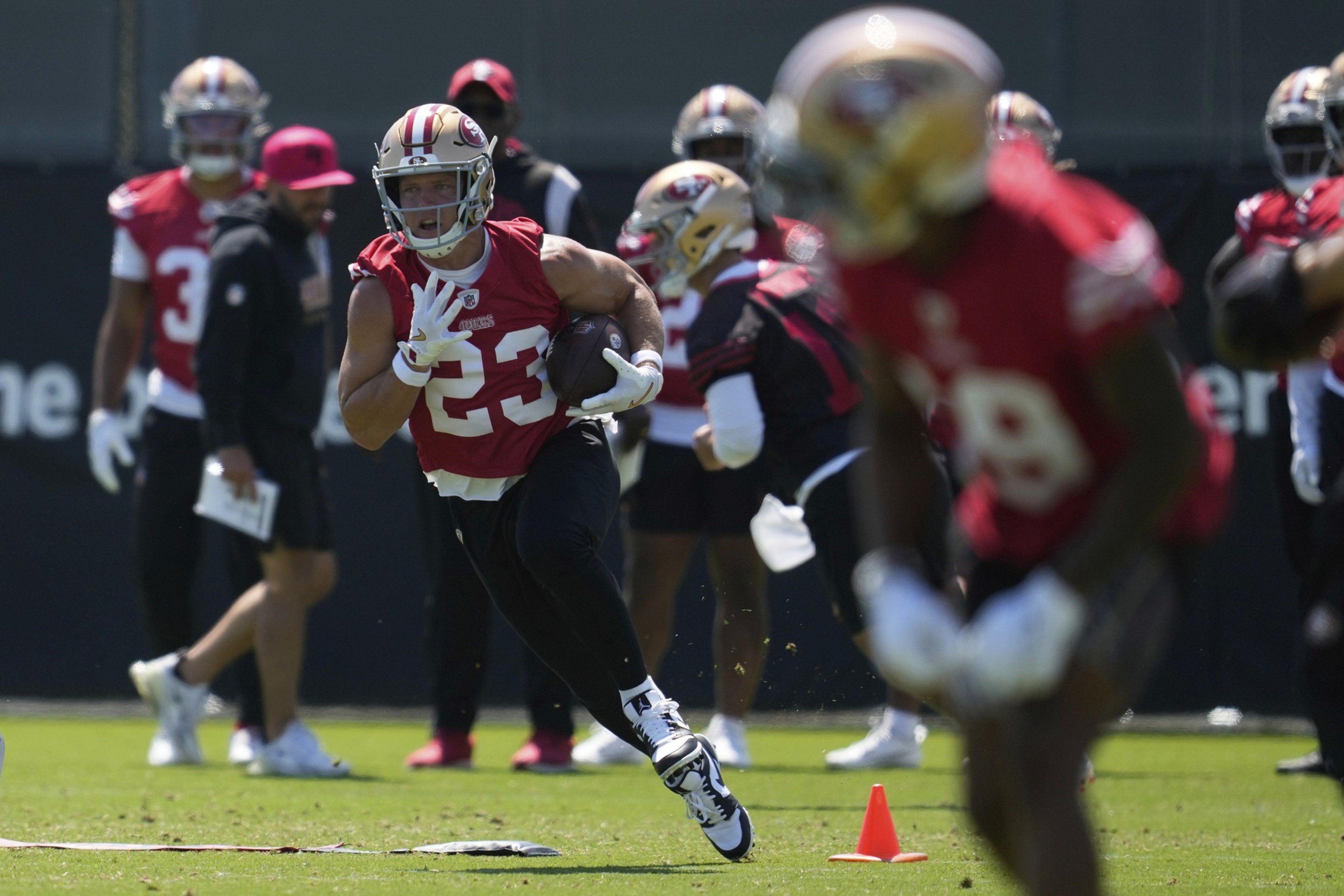 A football player in red uniform and helmet, holding a ball, is running on a green field. Other players and coaches in red are visible in the background.