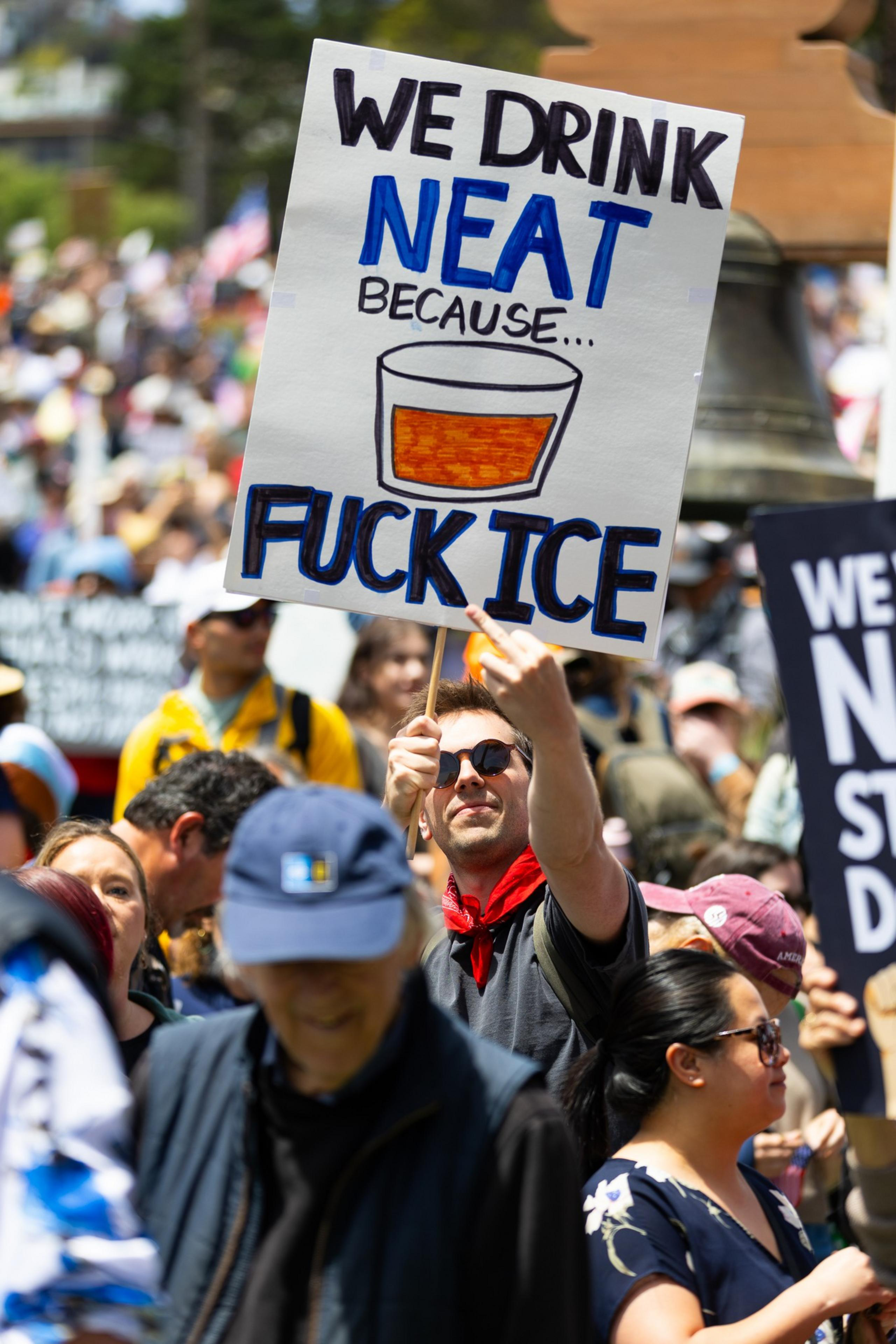 A person holds a sign reading &quot;WE DRINK NEAT BECAUSE... [EXPLETIVE] ICE&quot; at a crowded outdoor protest, with a sketch of a drink and people around them.