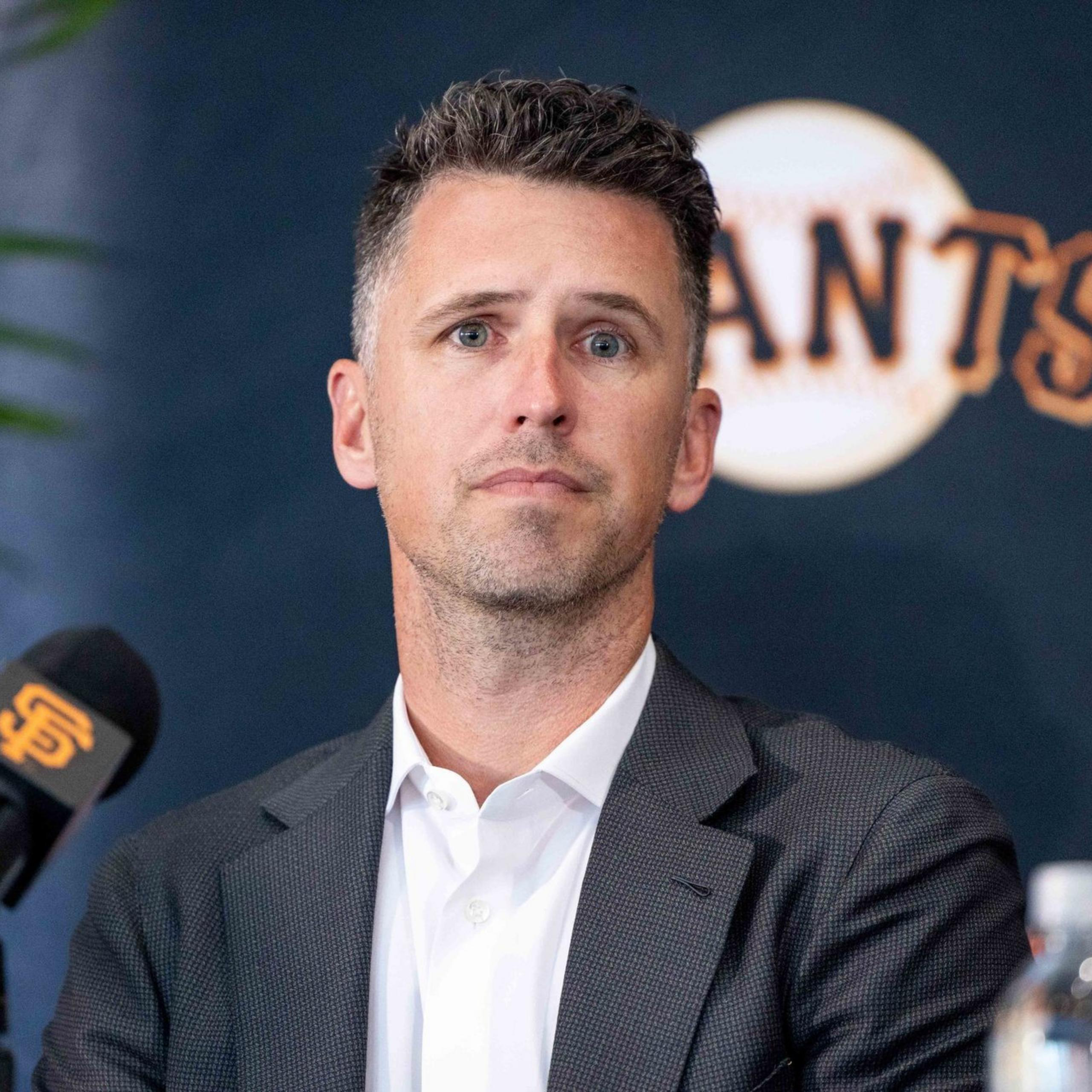 A person in a suit sits at a press conference with a mic displaying the San Francisco Giants logo. The backdrop shows the team's name partially visible.