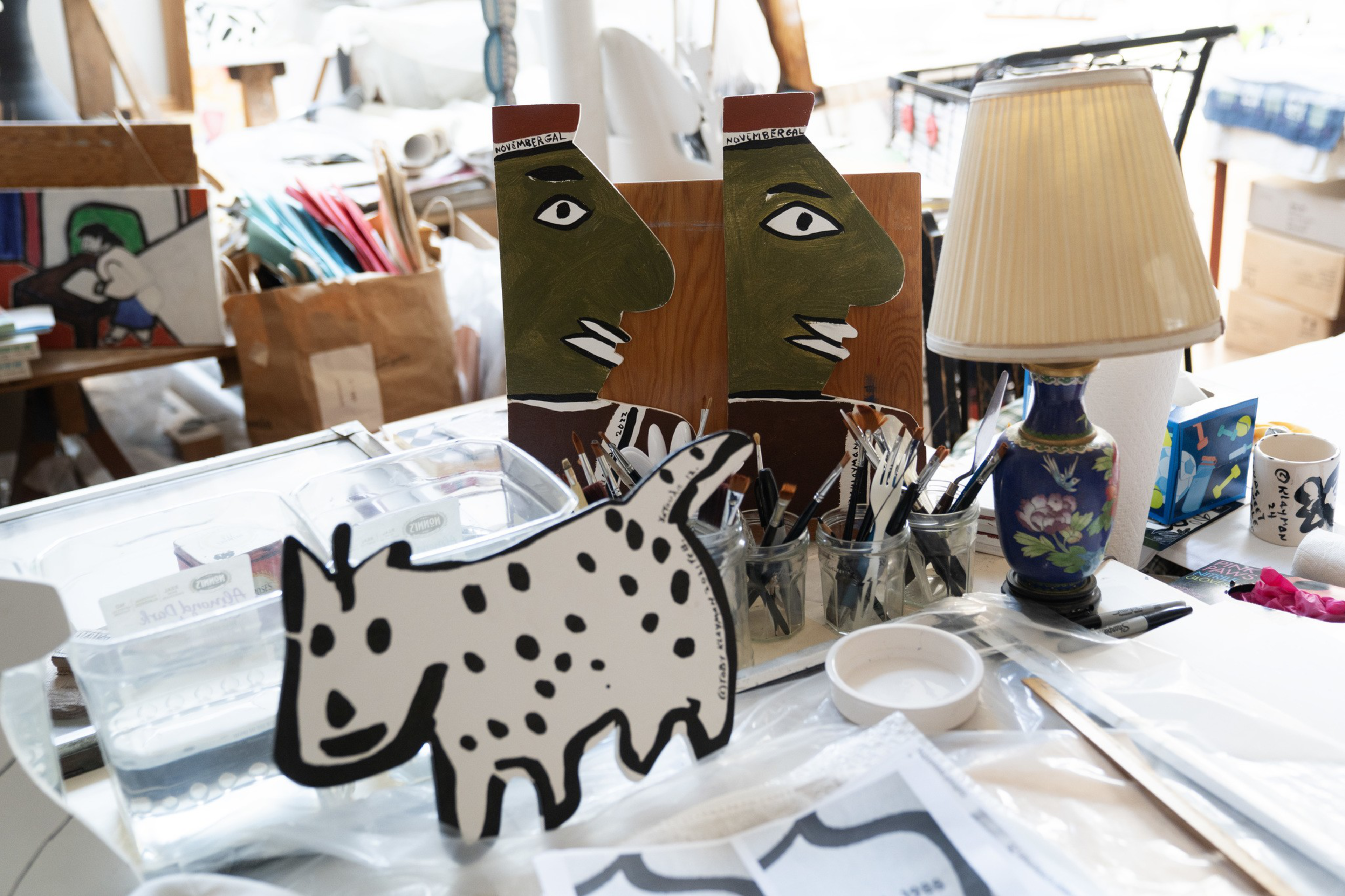 Various art supplies, including paintbrushes, a decorative lamp, and cutouts of a cartoon-like dog and two stylized human faces, are arranged on a cluttered table.