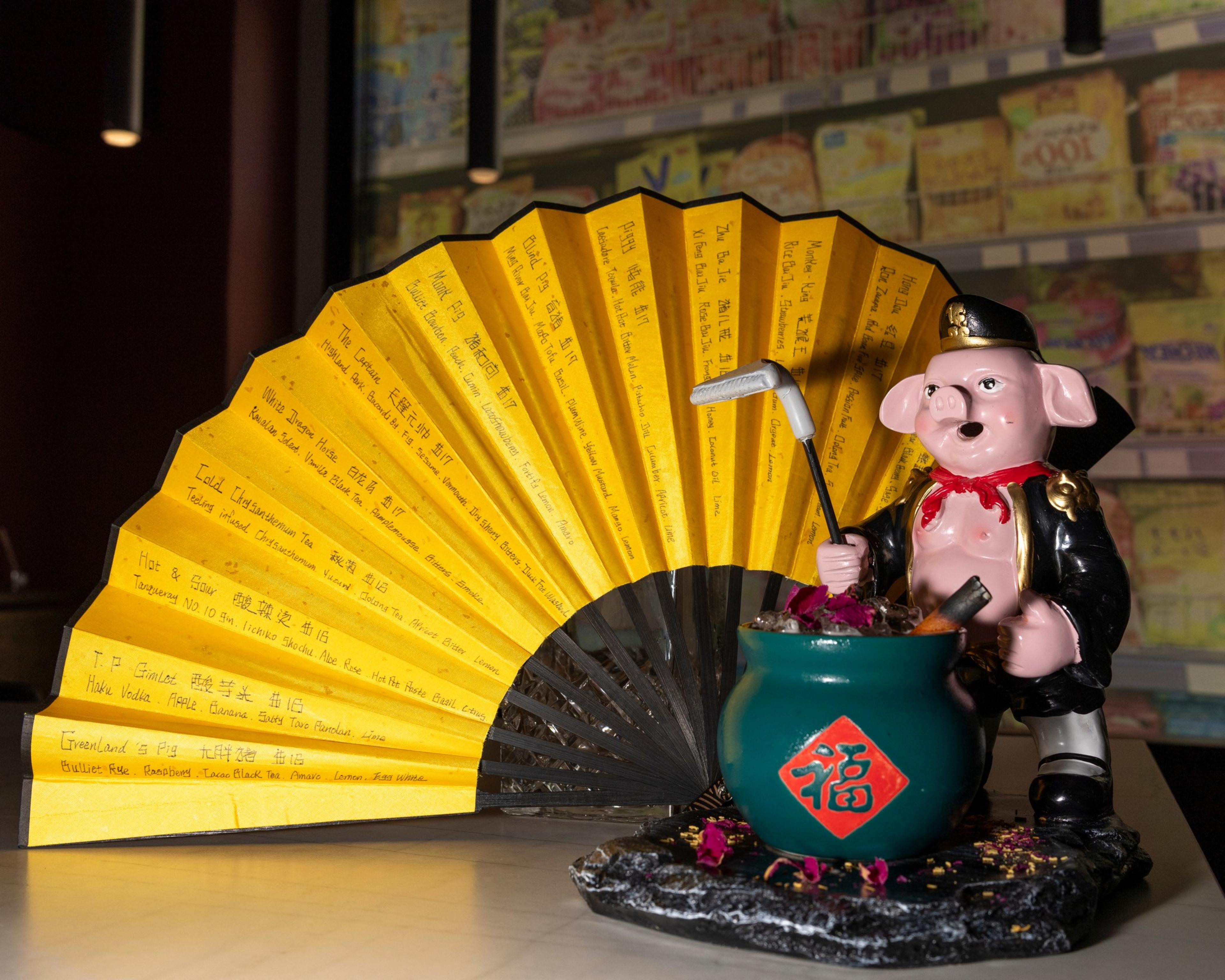The image features a pink pig statue in a suit, holding a golf club and standing next to a green pot with Chinese characters. A large, yellow fan with writing in the background.