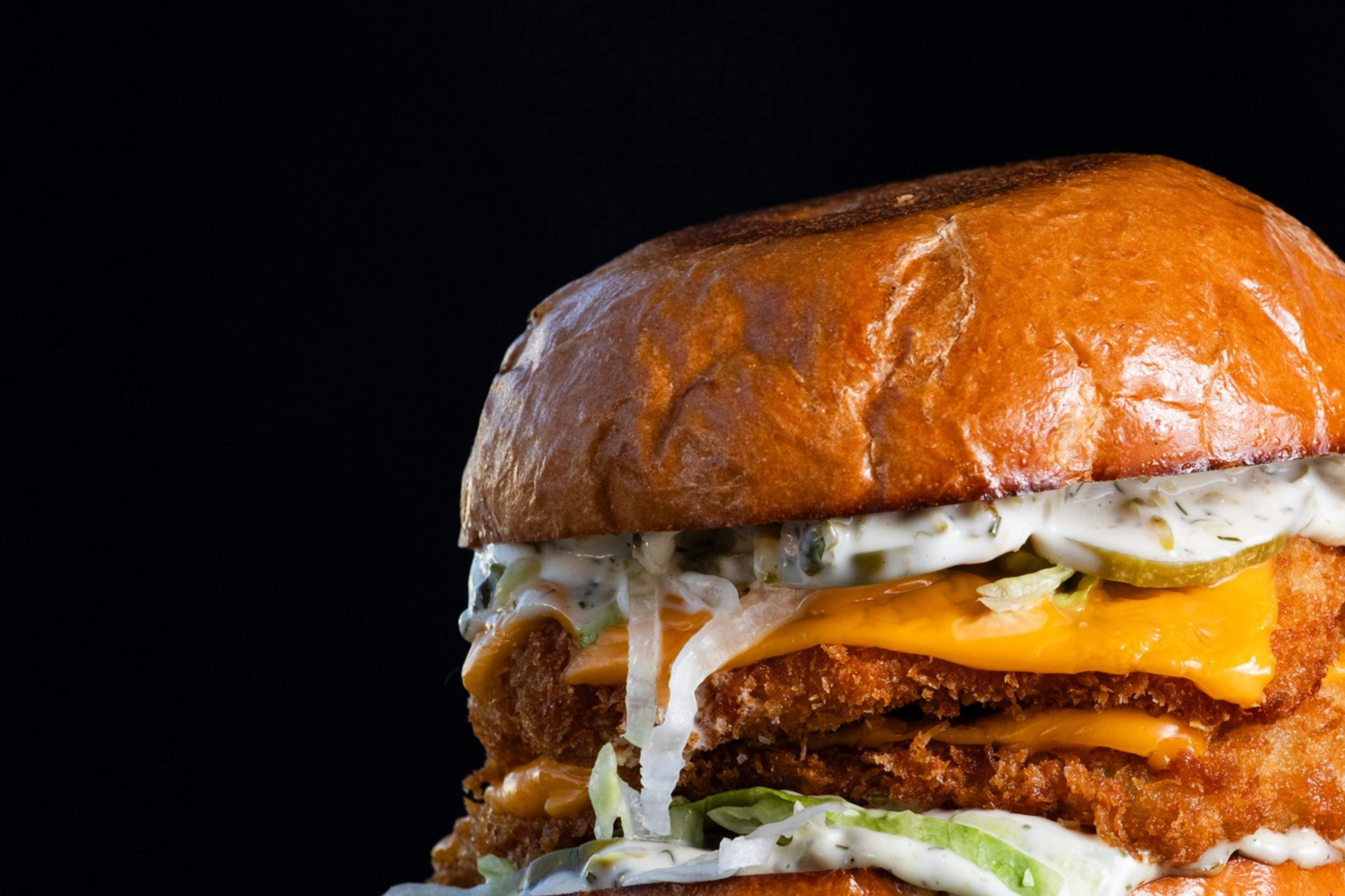 A crispy chicken sandwich with melted cheese, pickles, and creamy sauce is layered between a shiny, toasted bun on a plate against a dark background.