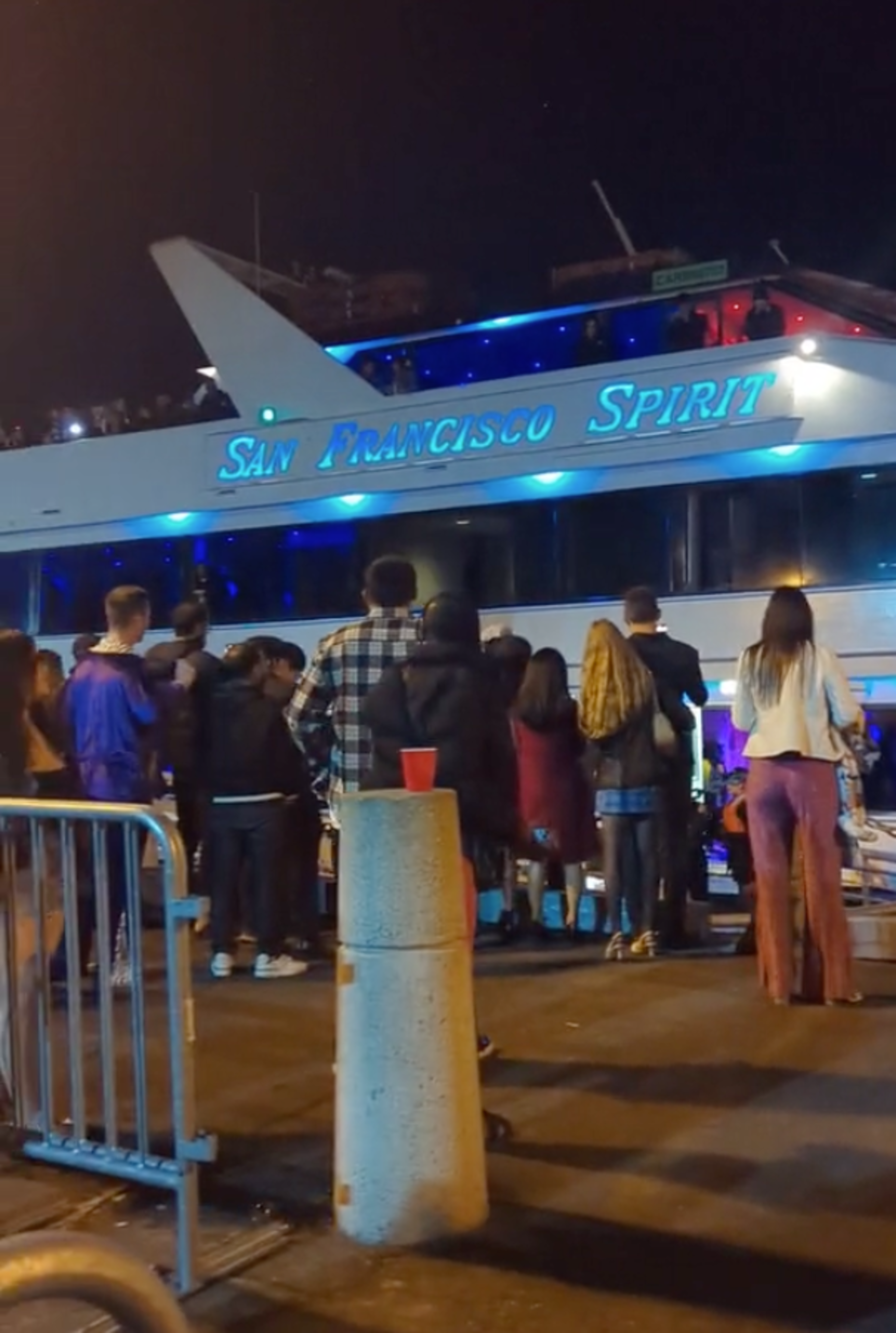 A crowd stands near a docked ship named "San Francisco Spirit" at night, with blue lights illuminating the vessel. A red cup is on a nearby concrete bollard.