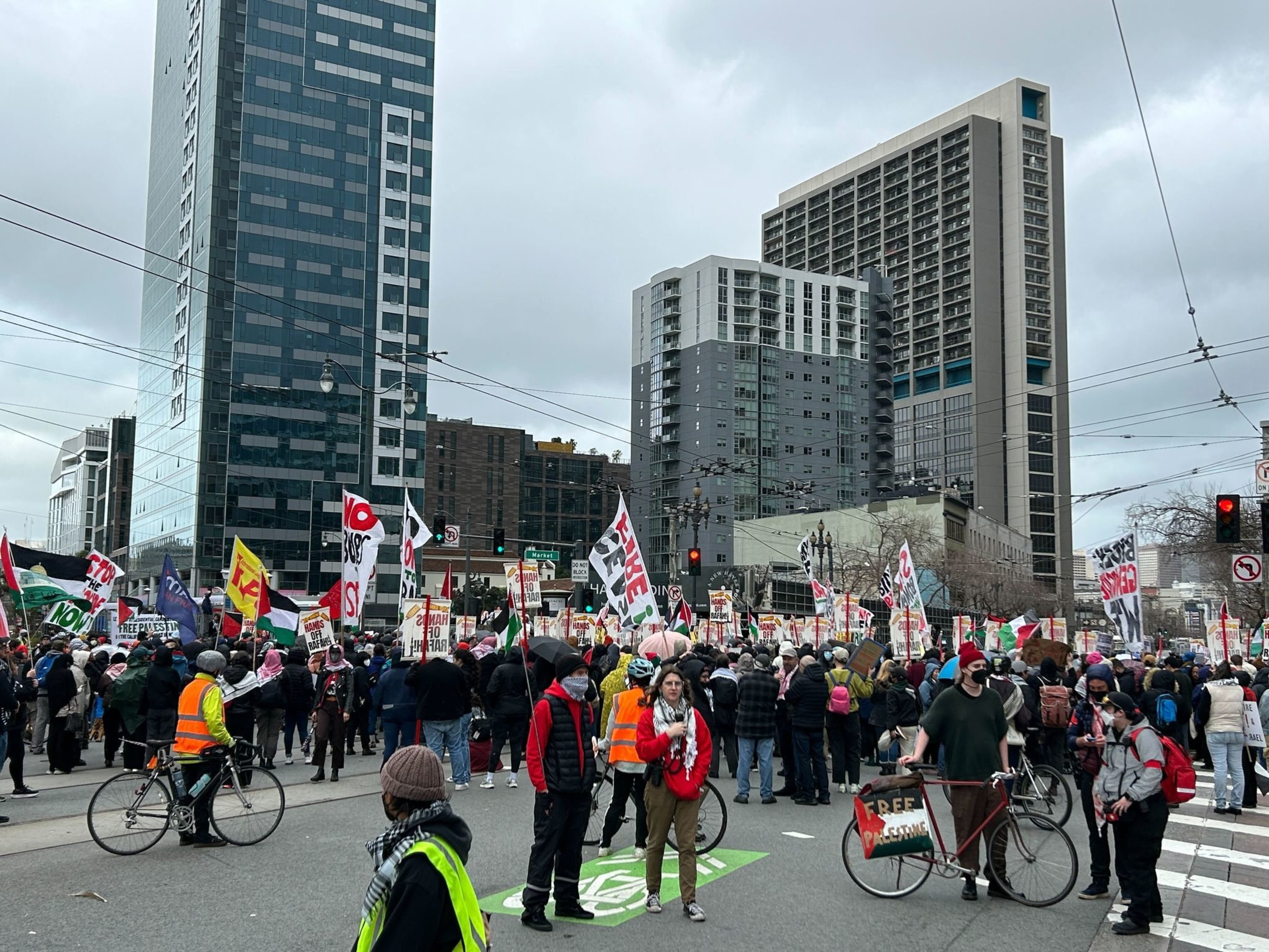 A crowd of protesters hold signs and fill a city intersection under mostly cloudy skies.