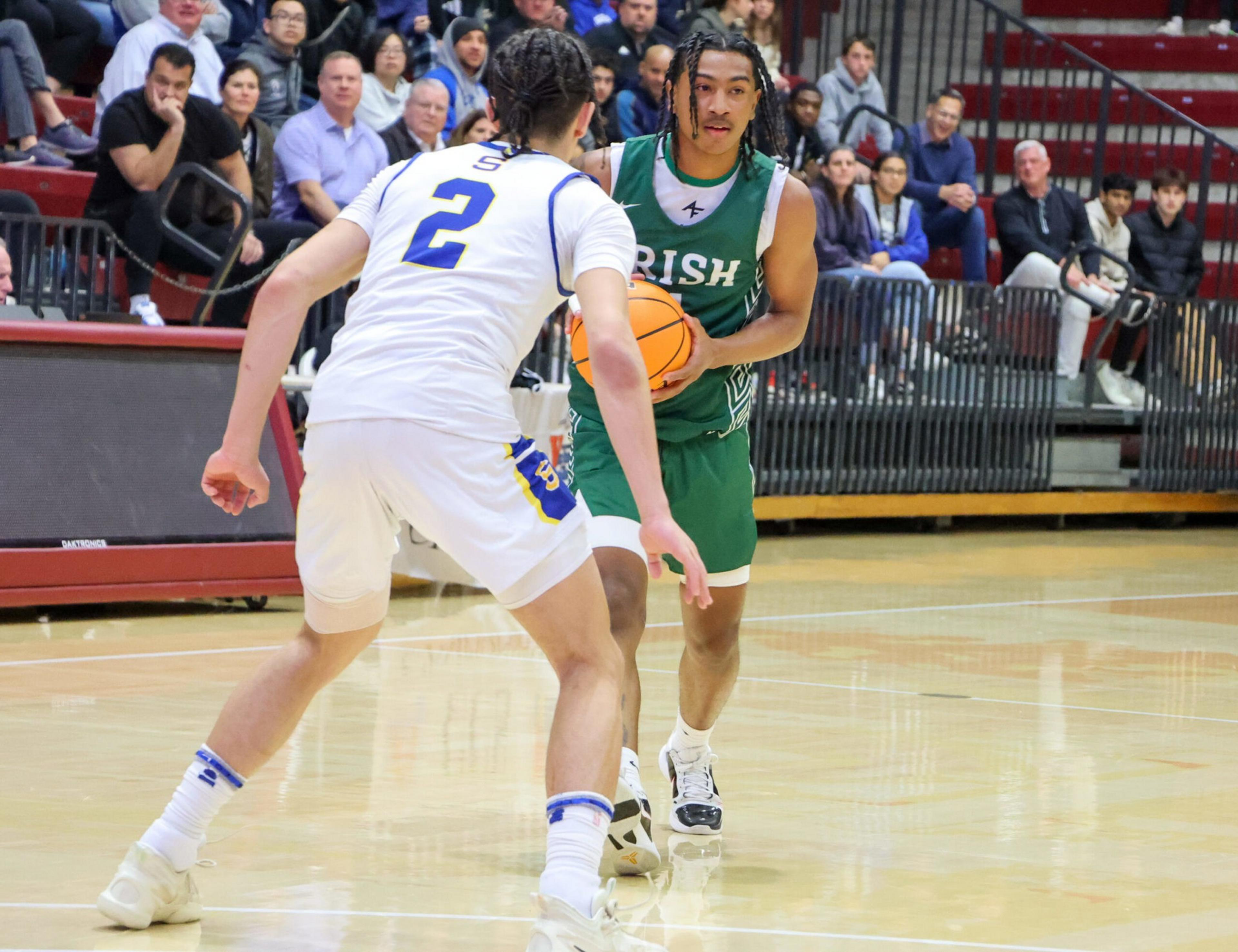 A basketball player in a green "Irish" jersey dribbles while being closely defended by a player in a white jersey numbered 2, with spectators watching.