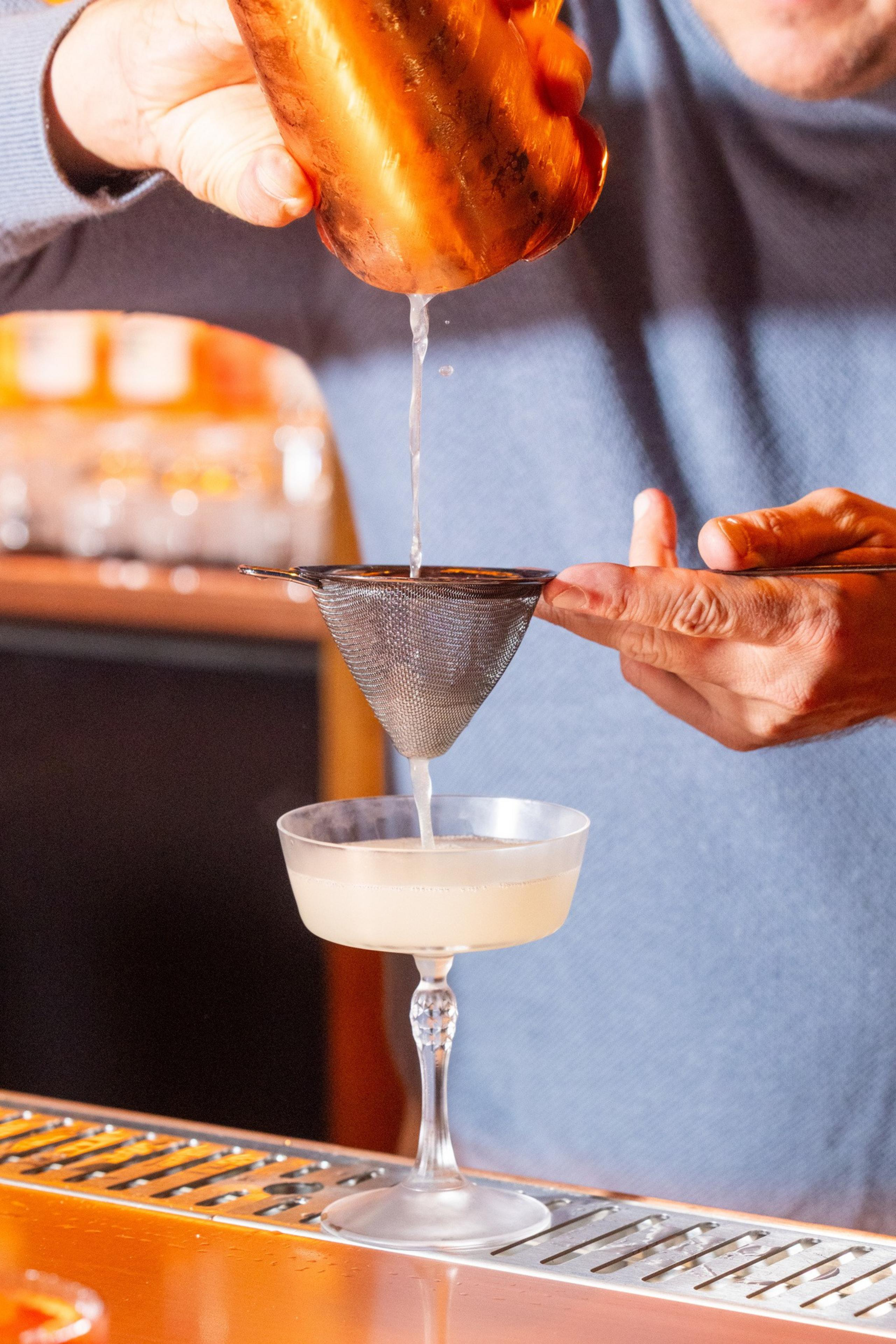 A person strains a cocktail from a copper shaker through a fine mesh strainer into a coupe glass on a bar counter.