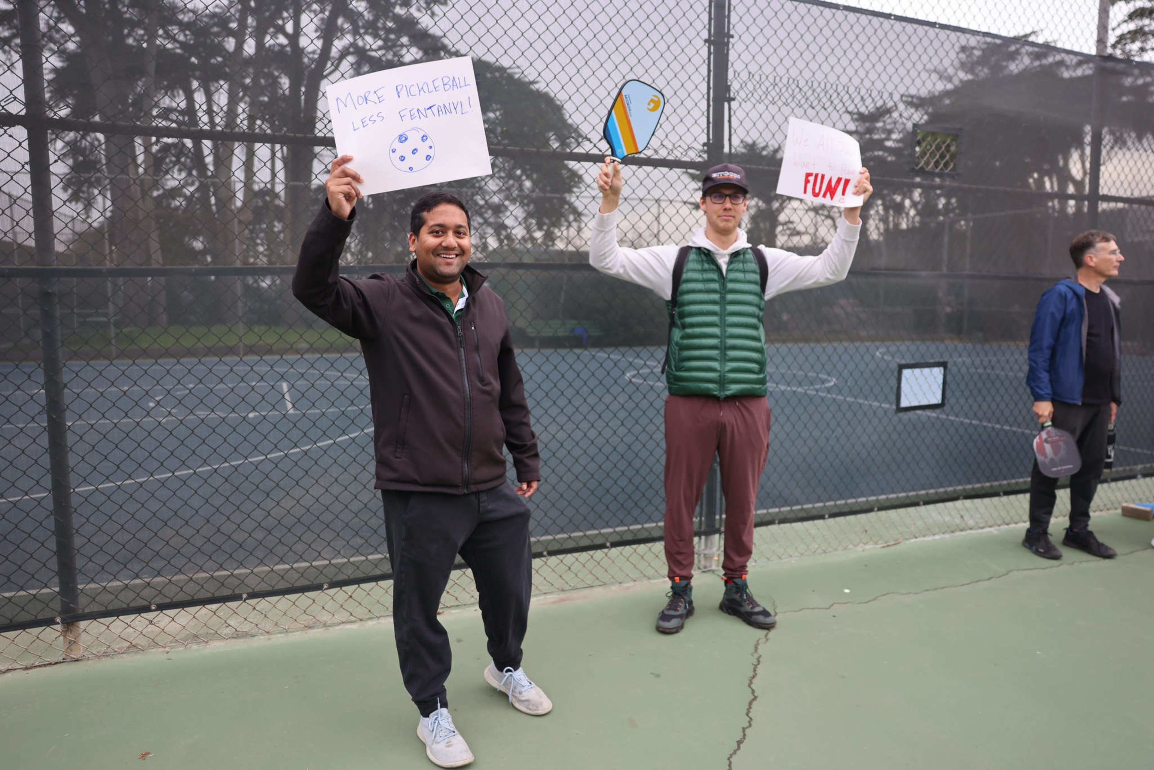 Two men stand on a tennis court, holding signs with "Make Pickleball Less Excruciating" and "FUN!" written on them.
