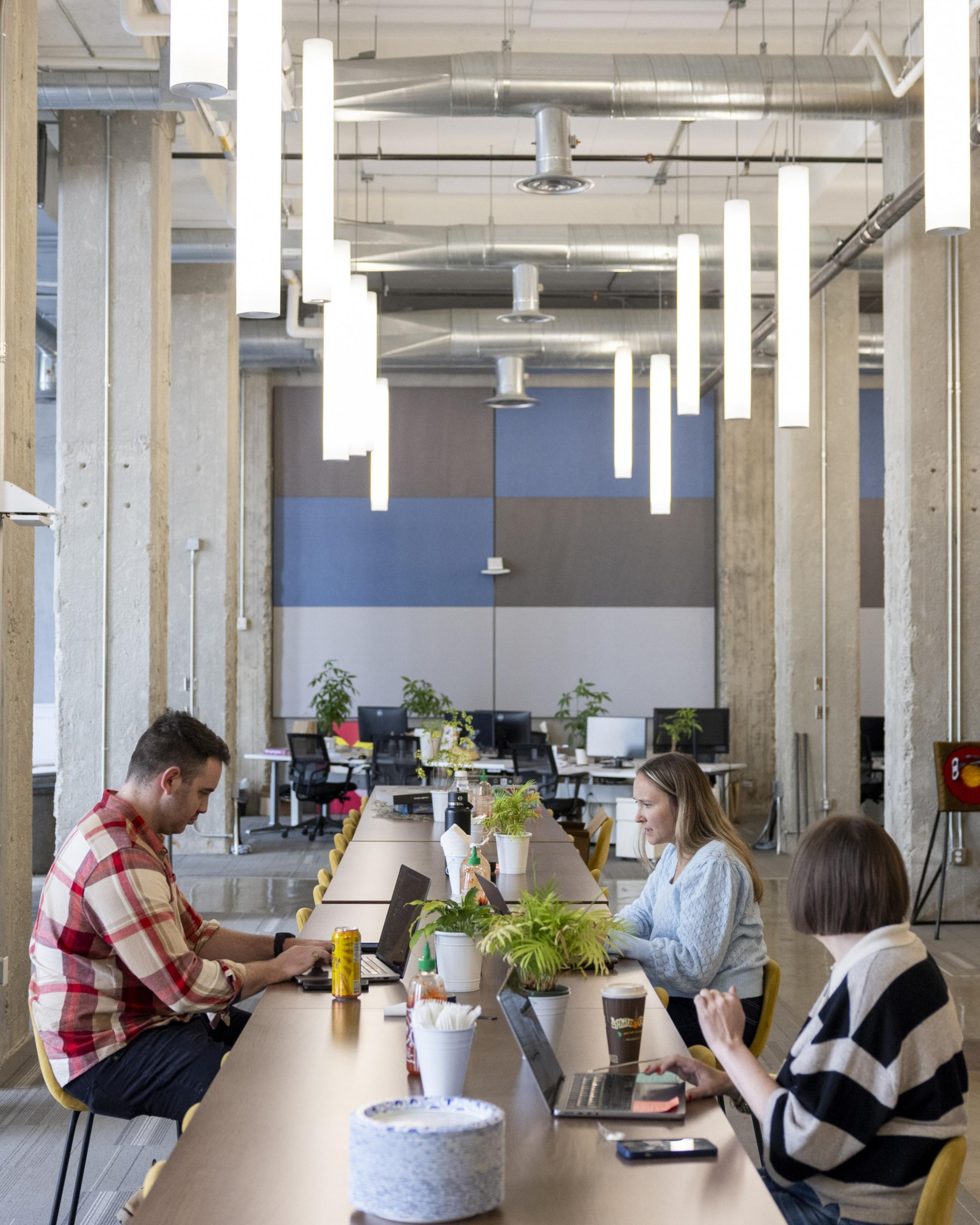 Three people work on laptops at a long communal table in a modern office with high ceilings, exposed ducts, concrete pillars, plants, and hanging lights.