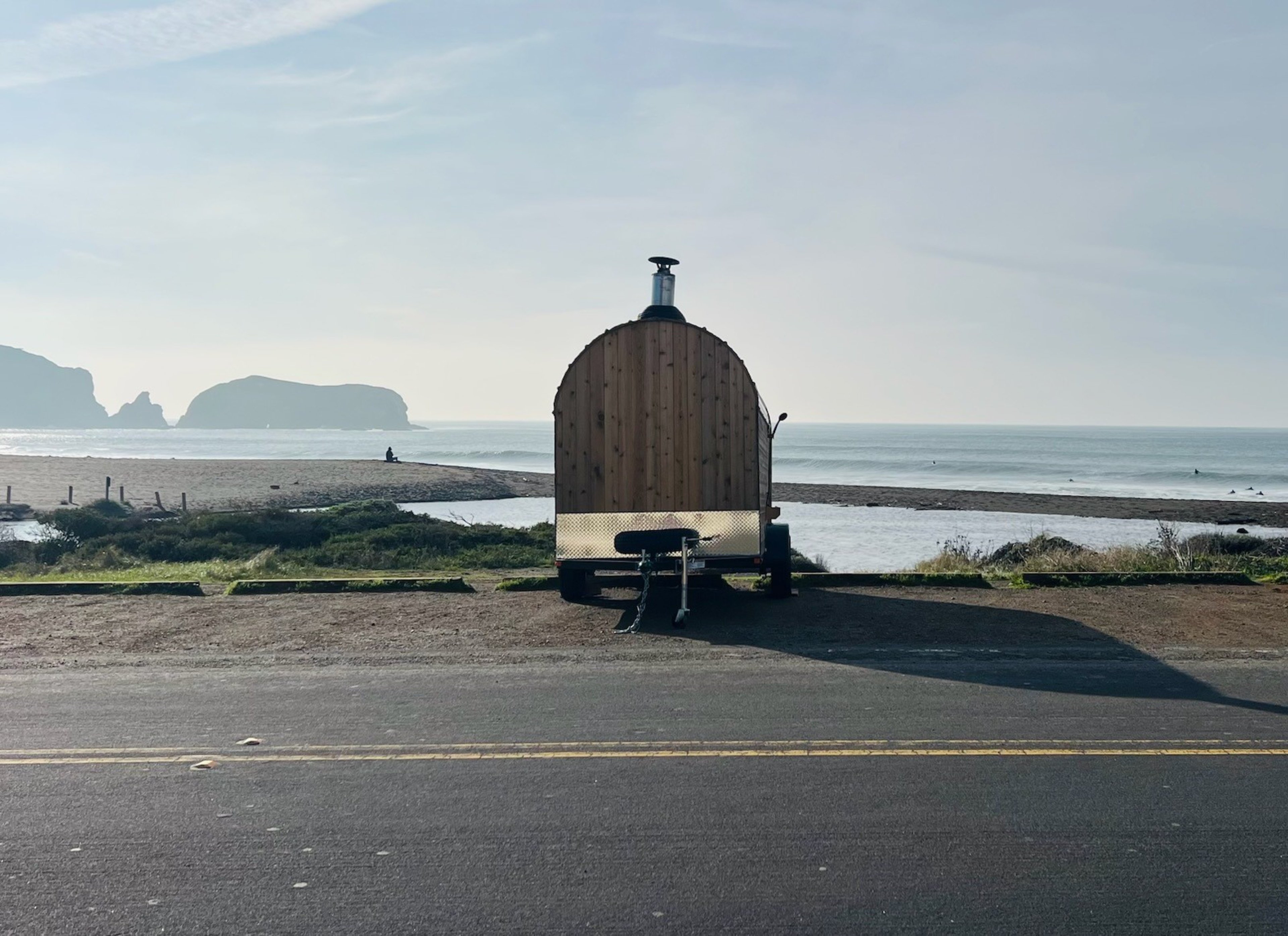 A small wooden trailer is parked by the ocean. Behind it, a sandy beach extends to the water, with rocky islands in the distance and a few people surfing.
