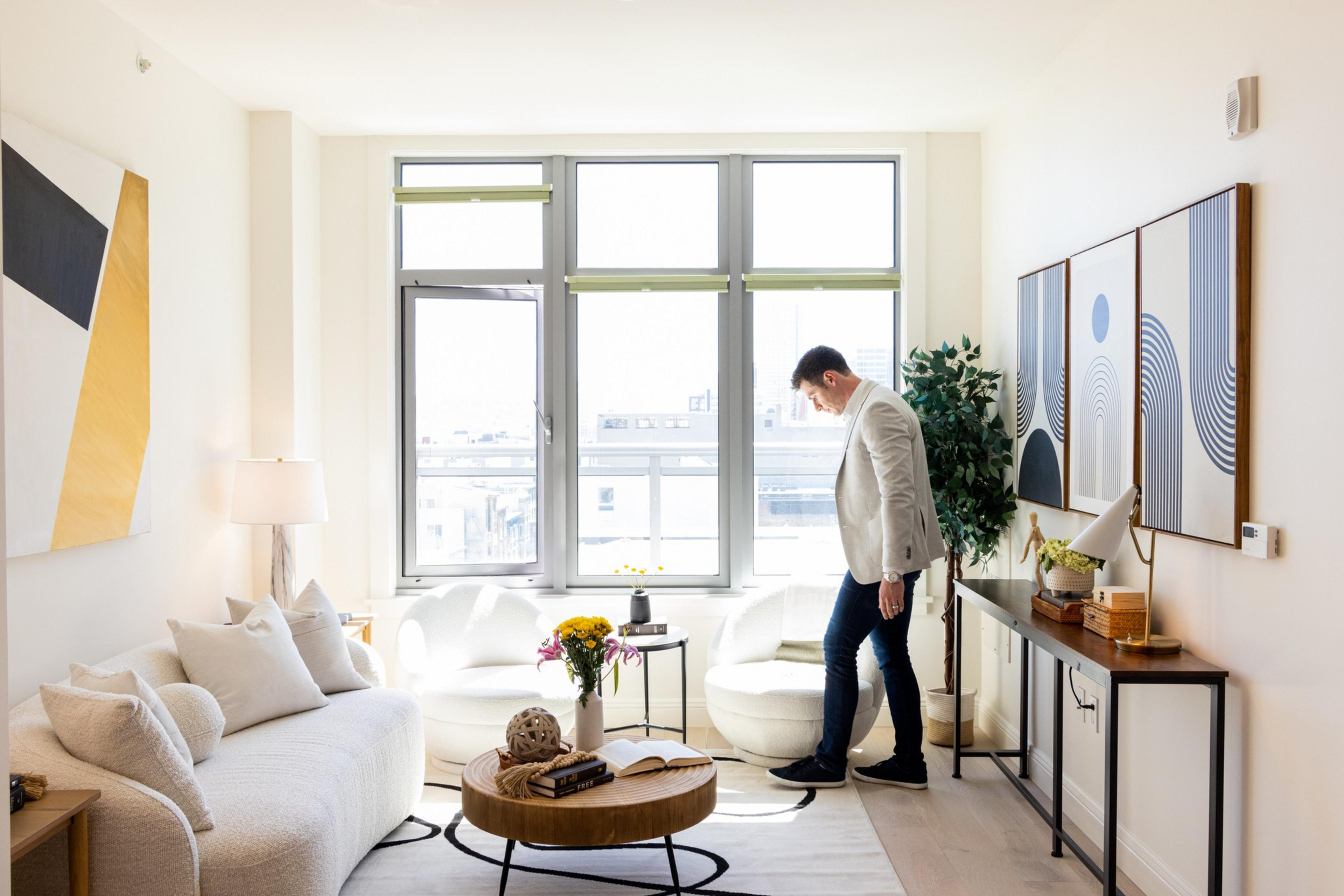 A tidy and brightly lit room with modern furniture features a man standing near a window. The room includes a sofa, round coffee table, large paintings, and houseplants.
