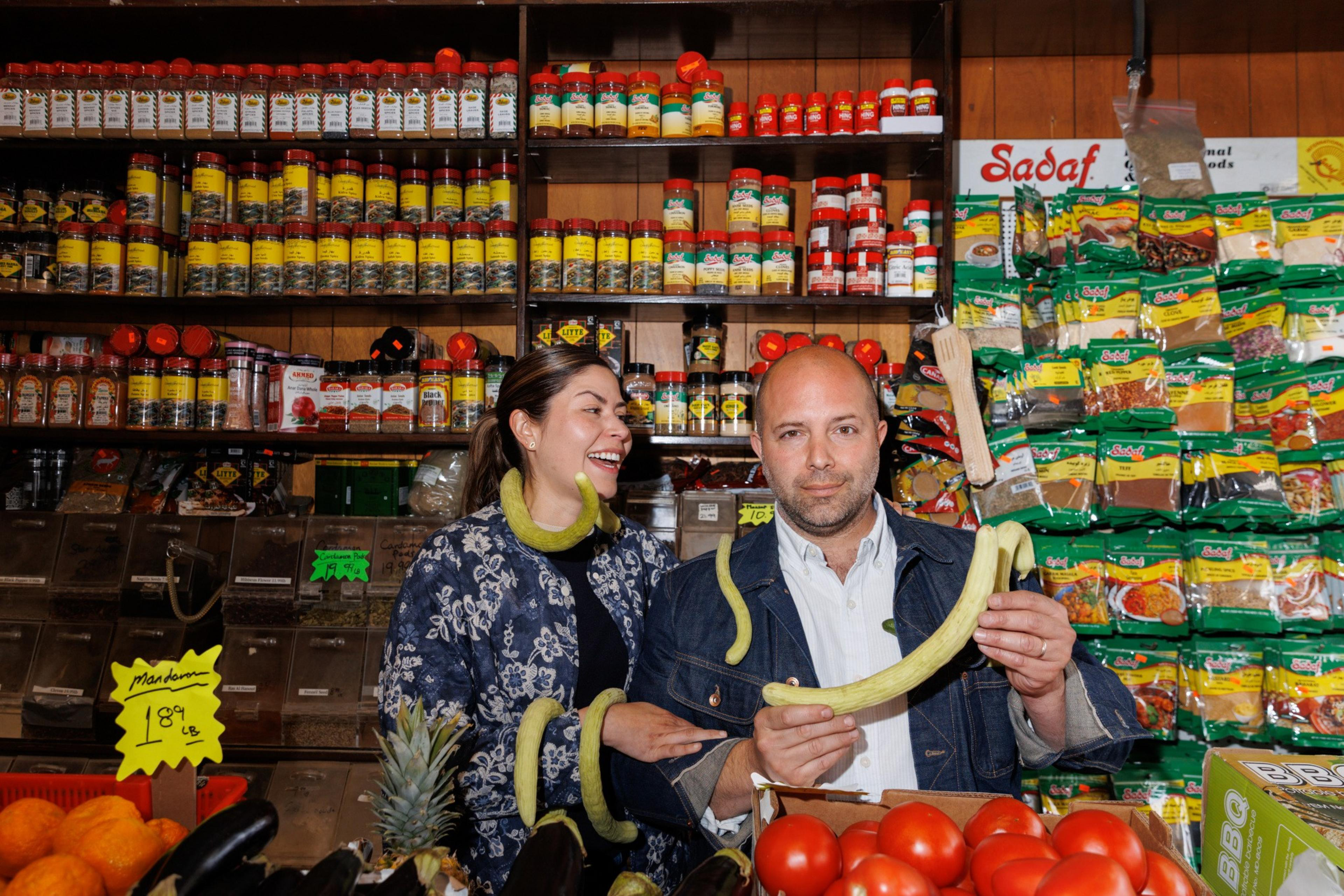A woman and a bald man stand in a colorful grocery store aisle with spices behind them. The man holds a long vegetable, and both have playful expressions.