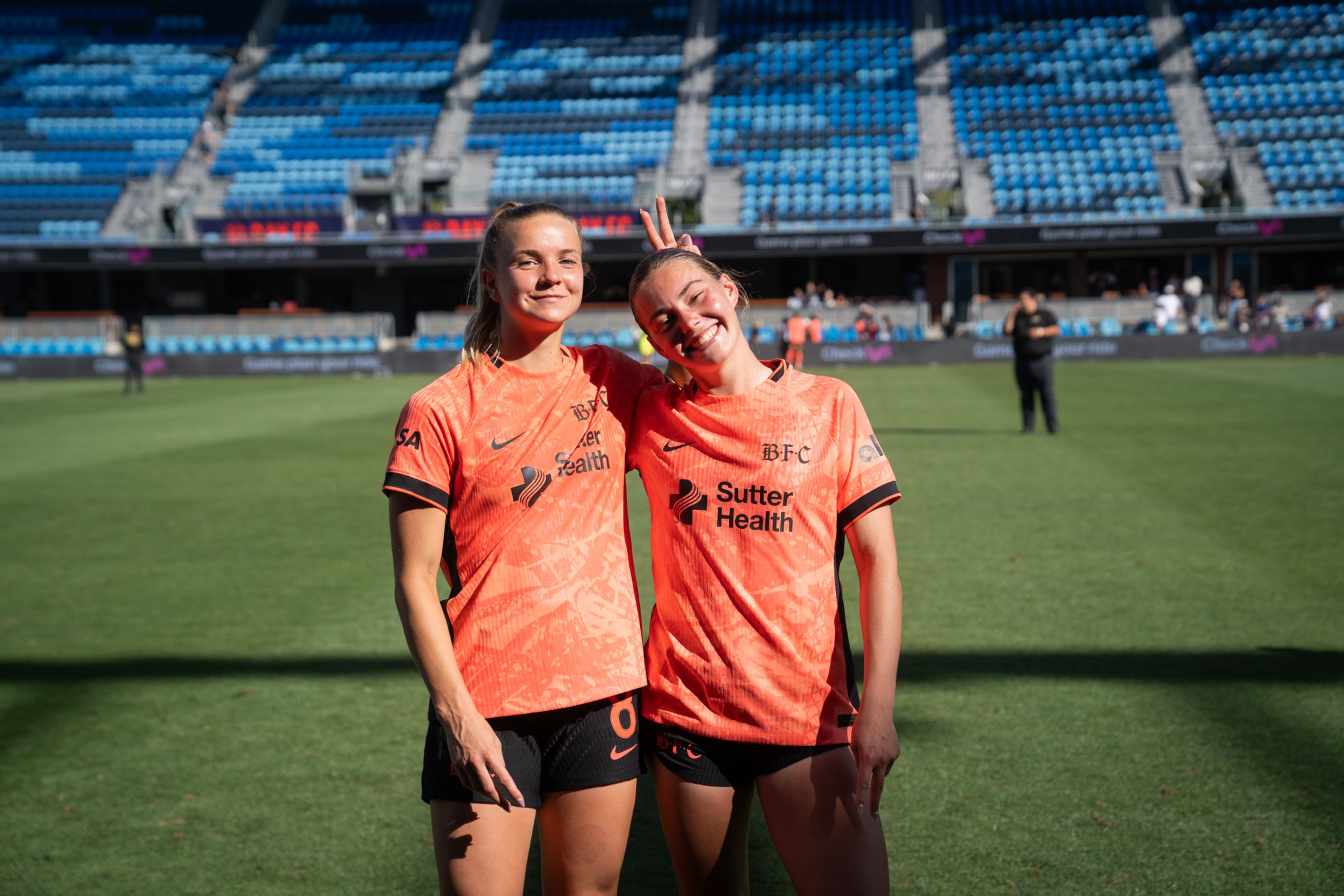 Two female soccer players in orange jerseys stand on a field, smiling, with one playfully giving bunny ears to the other in an empty stadium.